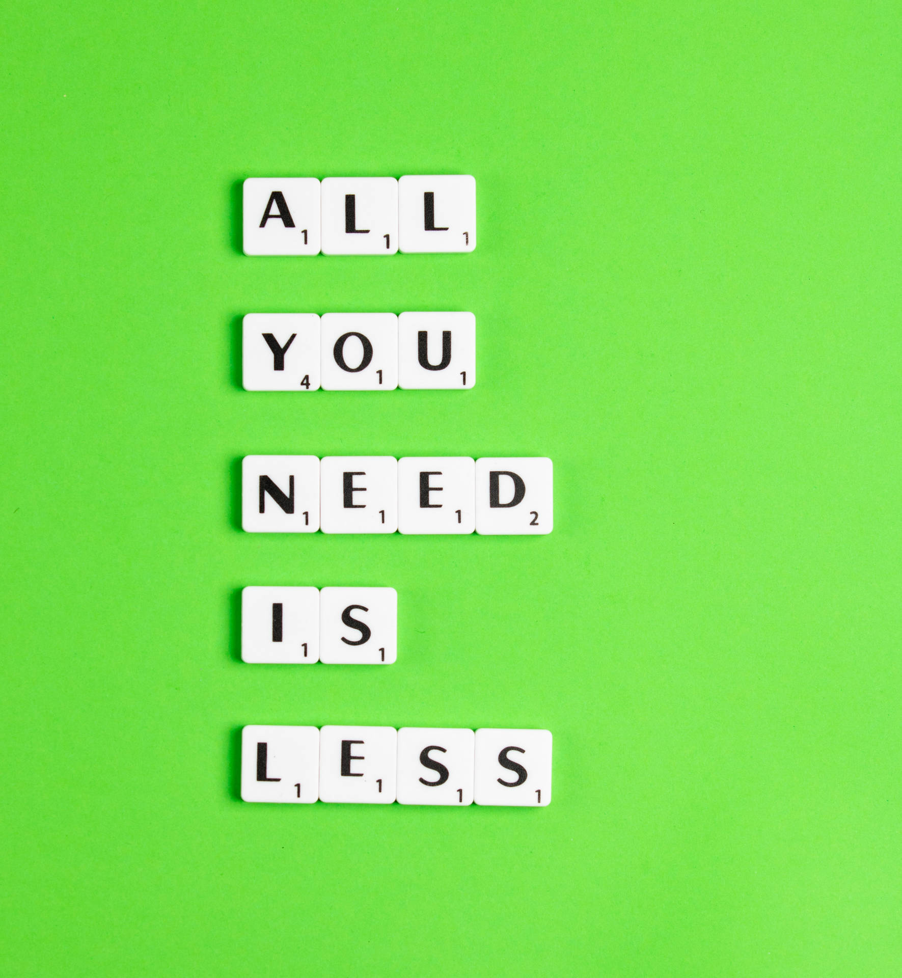 白色 Scrabble 字母磚垂直排列，在亮綠色表面上拼出 'All You Need Is Less'。