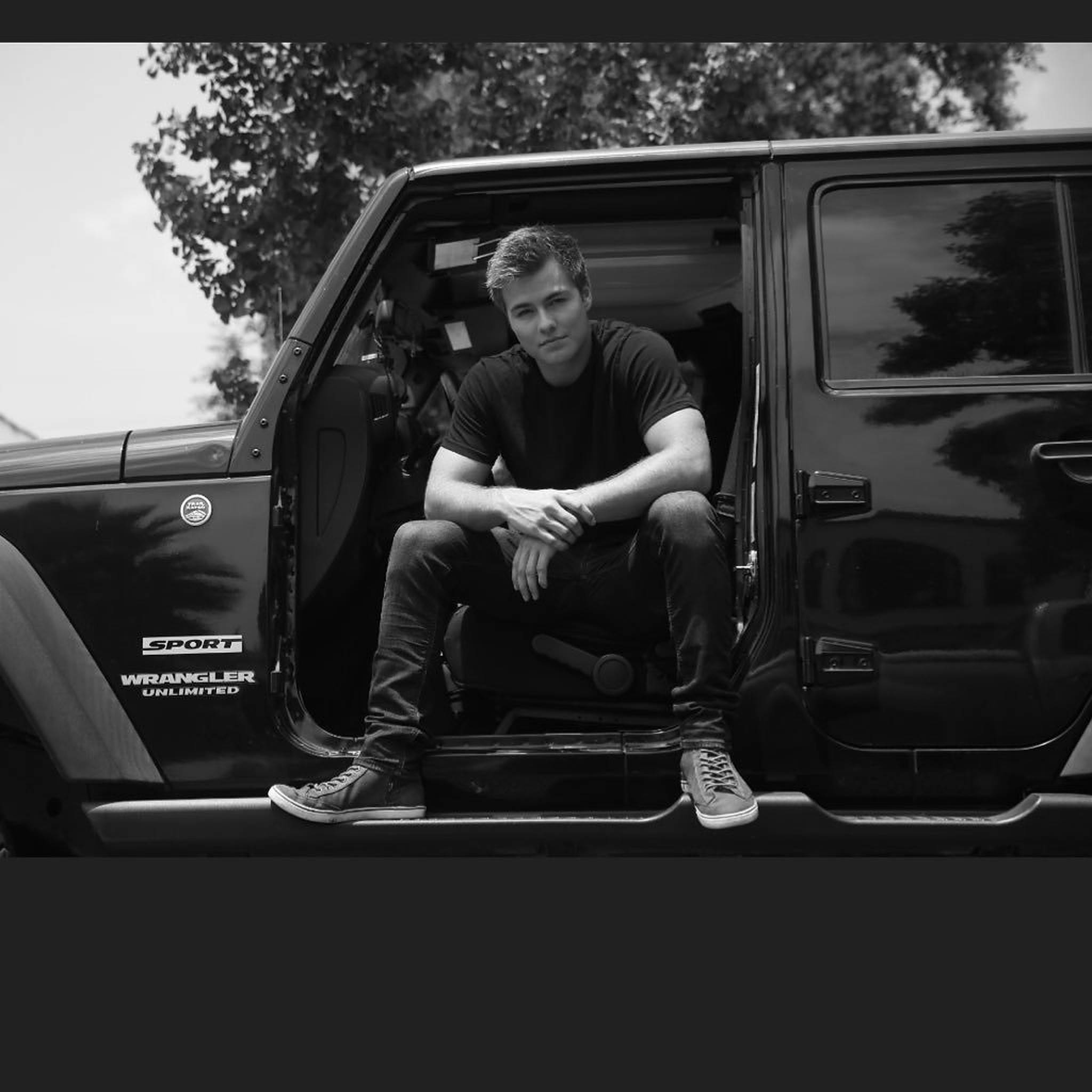 佩頓·梅耶黑色汽車壁紙 壁紙 — 一名男子隨意地坐在黑色Jeep Wrangler Unlimited的敞開車門中，背景是樹木和天空。
