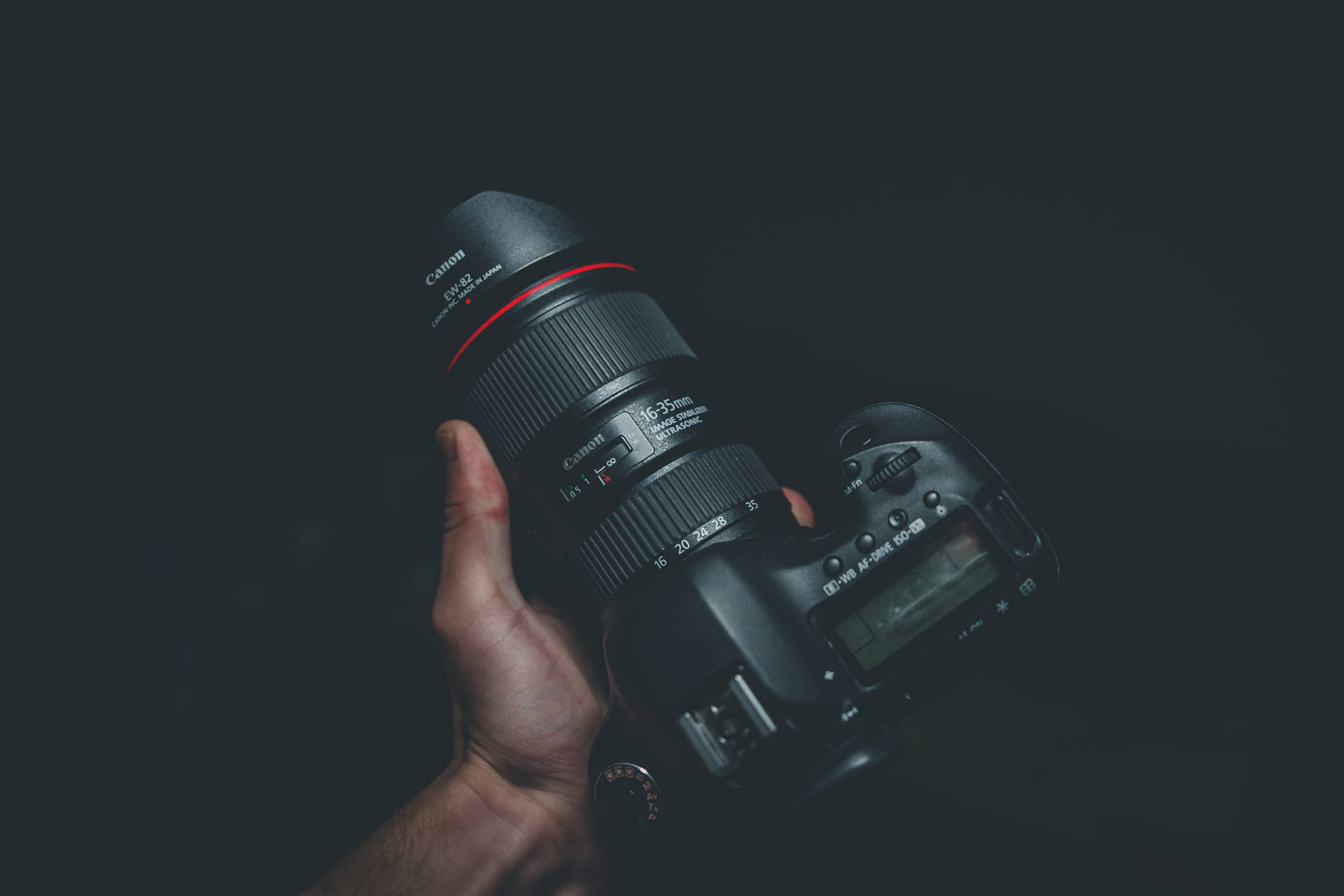 在黑暗環境中，一隻手拿著帶有紅色環形鏡頭的黑色Canon DSLR相機。