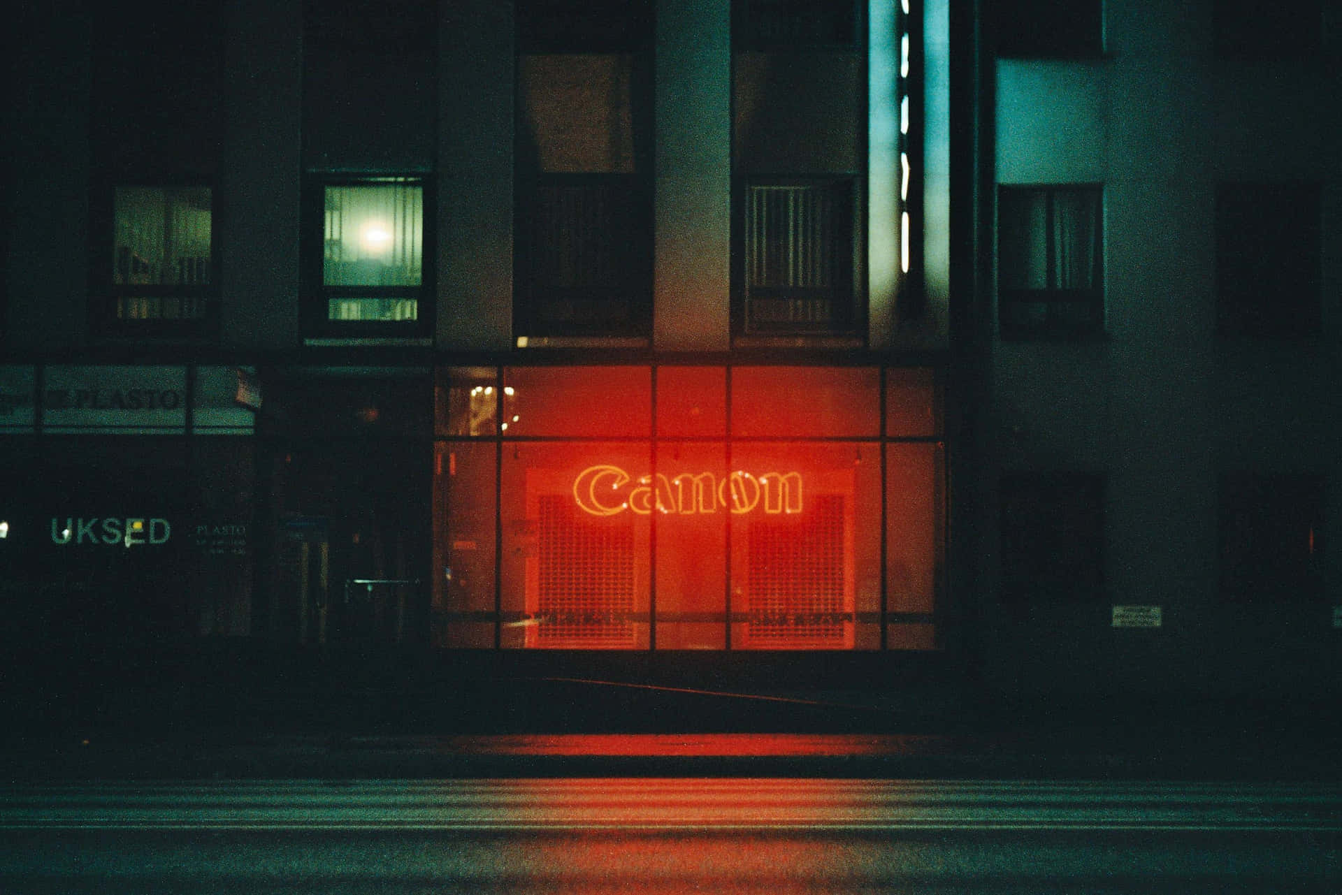 佳能霓虹燈 Tumblr 筆記本電腦壁紙 壁紙 — 一個發光的紅色Canon霓虹燈牌在夜間照亮濕漉漉的城市街道，在黑暗建築物外牆下的人行道上投射出倒影。