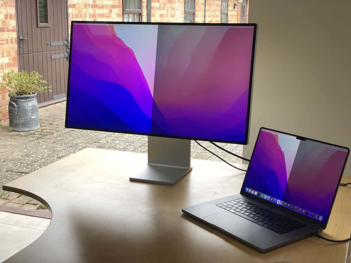 使用全能MacBook進行專案工作 壁紙 — 時尚的銀色顯示器和筆記型電腦在磚砌建築附近的木桌上展示充滿活力的紫色、藍色和粉紅色抽象桌布。