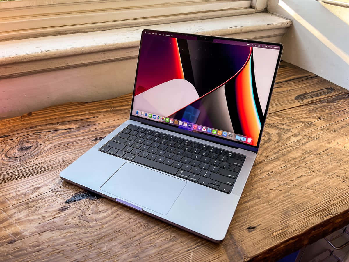 使用新款MacBook Pro更輕鬆完成線上工作 壁紙 — 一部銀色手提電腦，顯示色彩繽紛的抽象桌布，打開放在窗邊的鄉村風格木桌上。