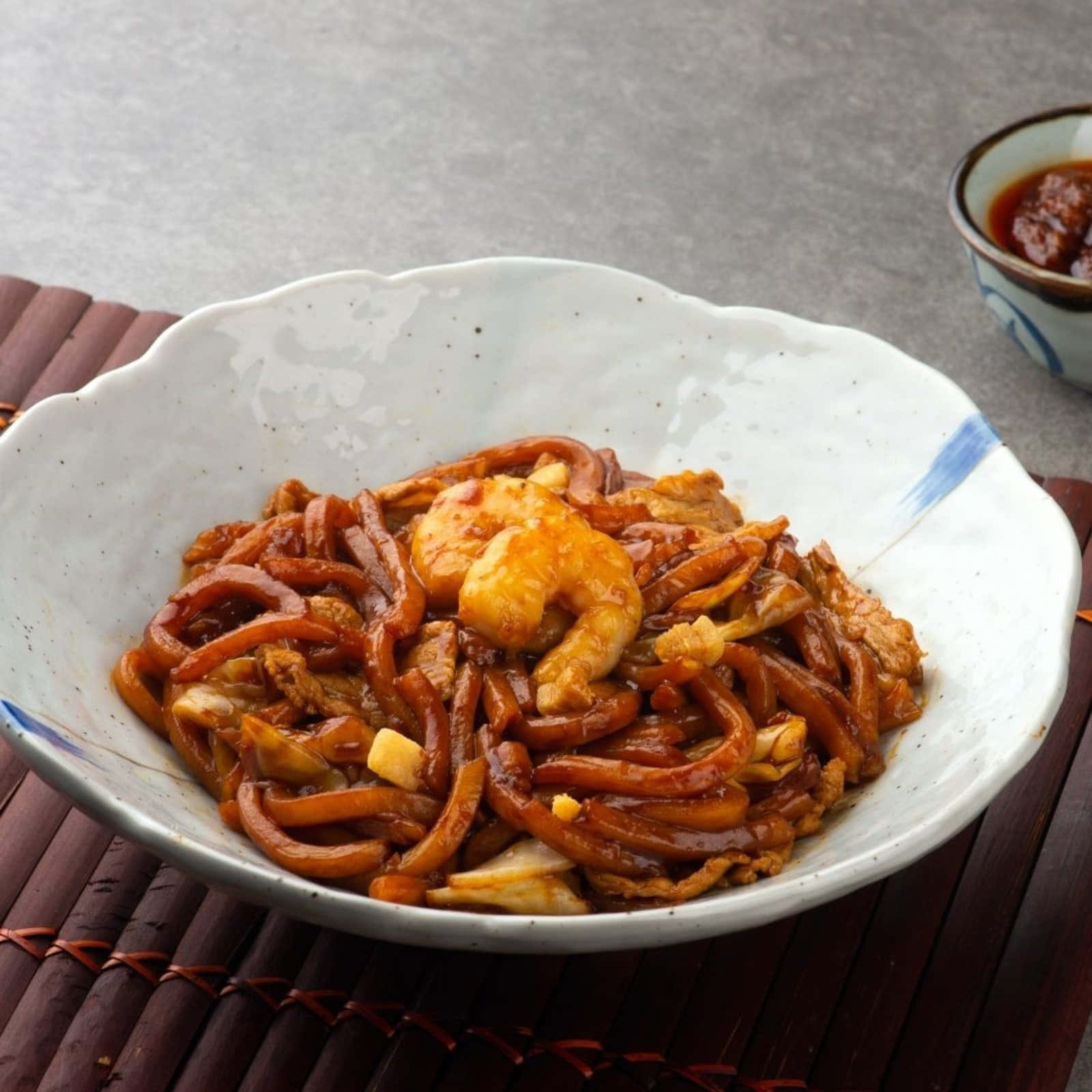 使用烏龍麵製作的福建麵 壁紙 — 一碗油亮亮的炒麵，配上蝦和蛋，放在竹蓆上，旁邊還有一小碗蘸醬。