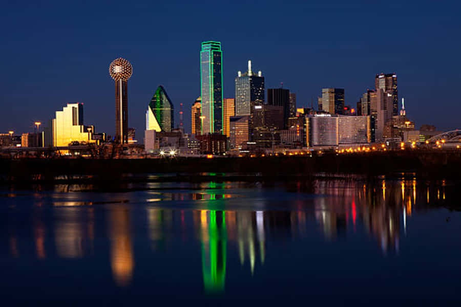 來探索美麗的德州風景吧 壁紙 — 達拉斯夜景，摩天大樓燈火輝煌，色彩繽紛的倒影在平靜的河面上閃爍。