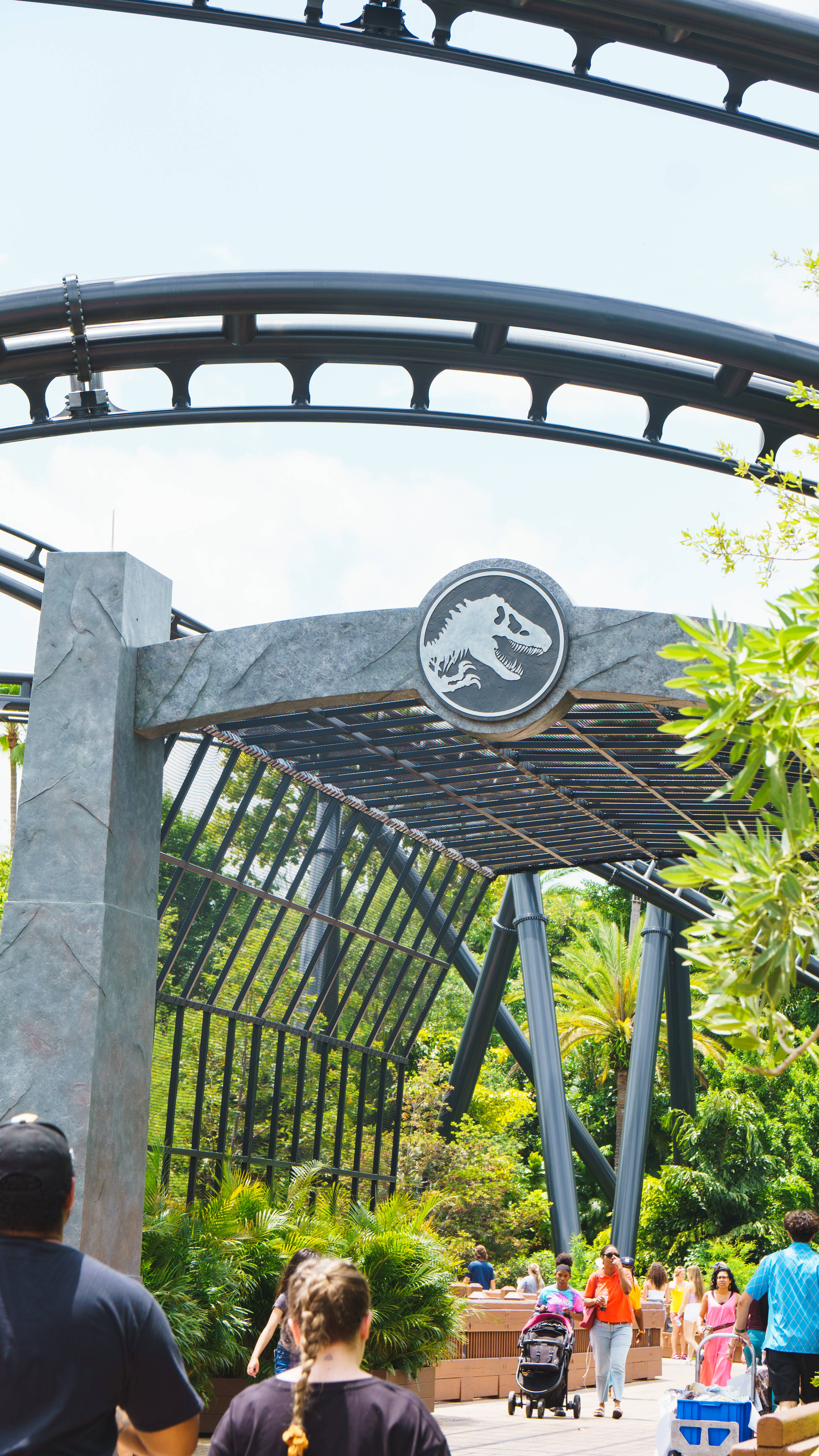 《侏羅紀世界》主題公園 壁紙 — 遊客在侏羅紀公園主題的過山車入口拱門下行走，拱門上有茂密的綠植和恐龍標誌。