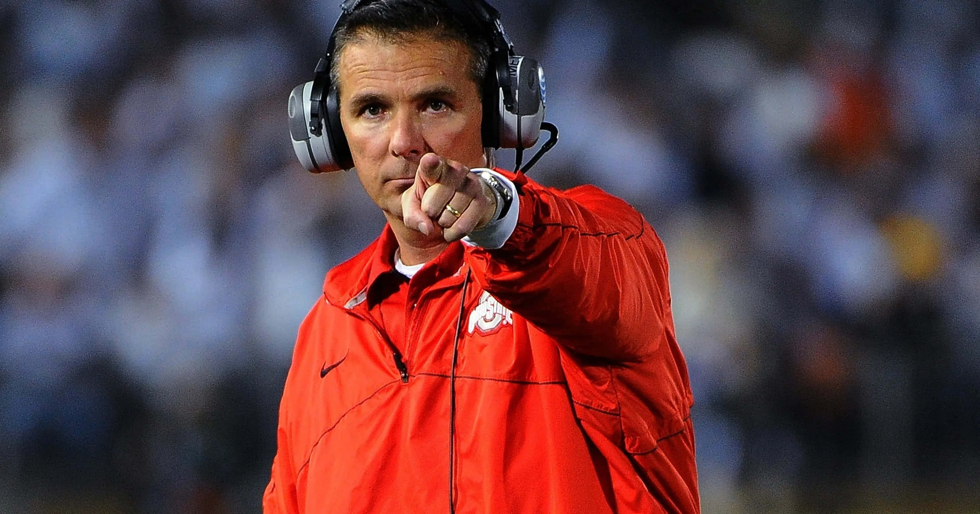 俄亥俄州立大學七葉樹隊教練 Urban Meyer 壁紙 — 一位專注的足球教練穿著紅色外套和耳機，在夜間比賽中在球場燈光下專注地指點。