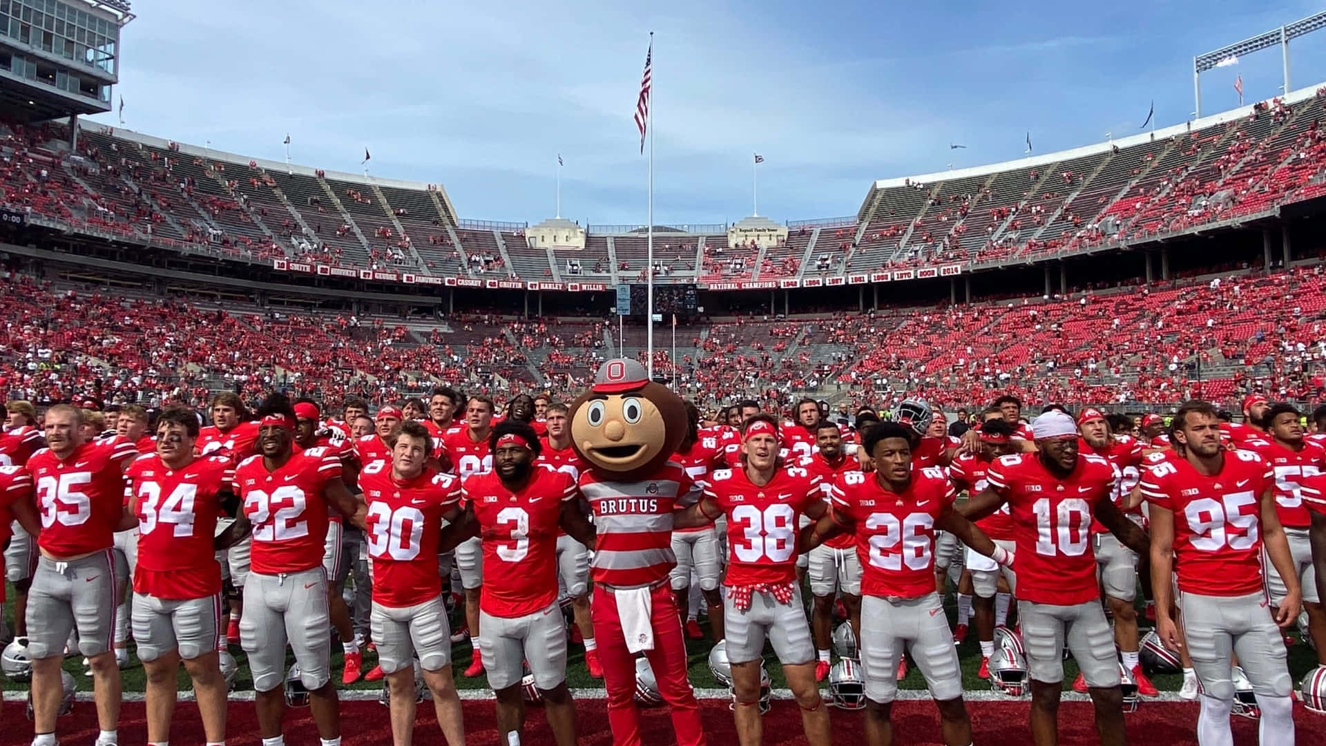 俄亥俄州 桌布 壁紙 — 俄亥俄州立大學美式足球隊與吉祥物Brutus the Buckeye在晴朗藍天下，一起站在座無虛席的球場上。