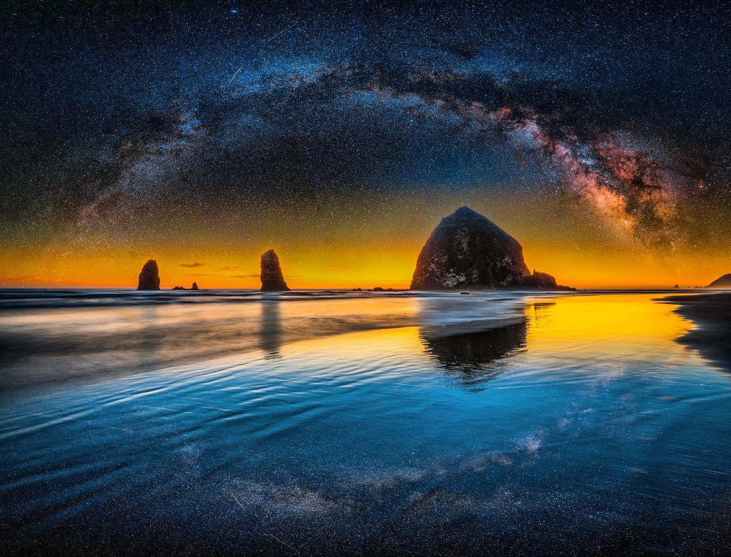 暮色中銀河在海蝕柱和大岩石之上，星空倒映在濕沙中。