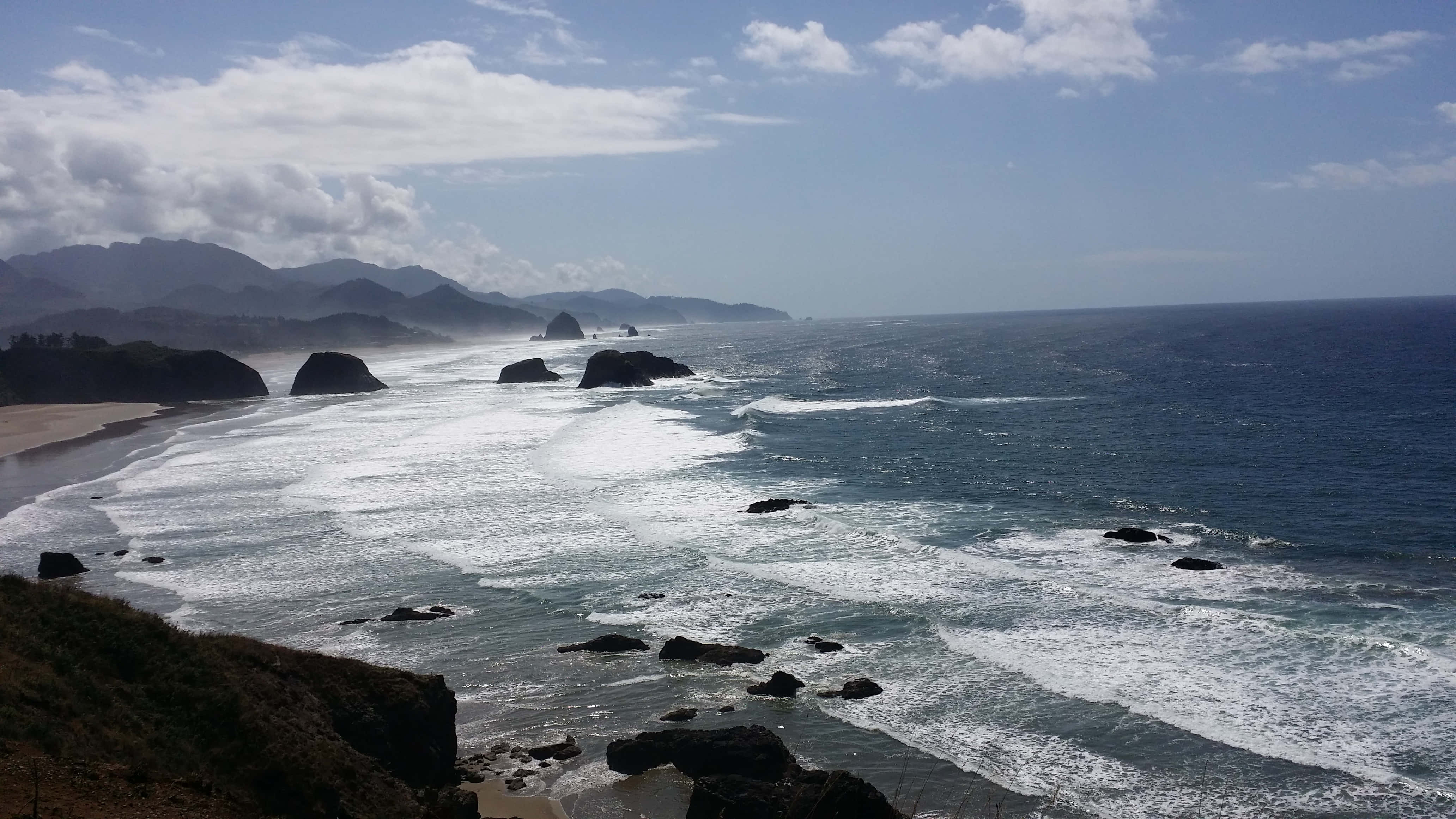 俄勒岡海岸的自然美景 壁紙 — 強勁的海浪拍打著崎嶇的黑色岩石，沿著蜿蜒的海岸線，天空部分多雲。