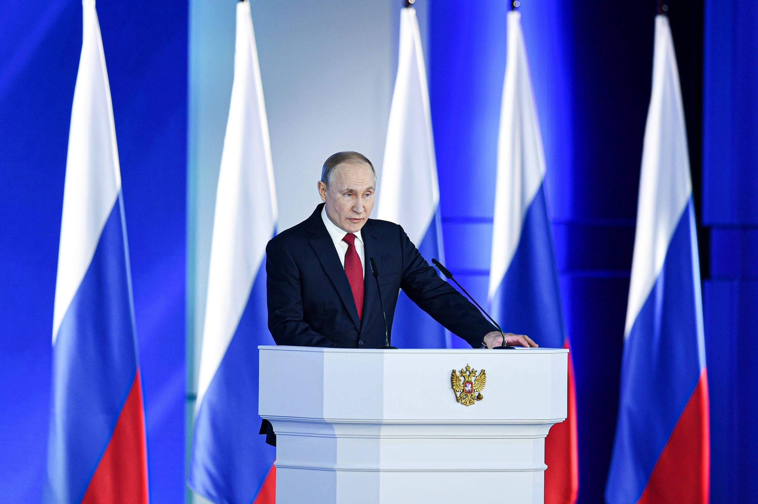 俄羅斯總統弗拉基米尔·普京於五國國旗前挺立 壁紙 — 一名身穿深色西裝、打紅色領帶的男子站在白色講台上，講台上有金色徽章，兩側是俄羅斯國旗，背景為藍色。