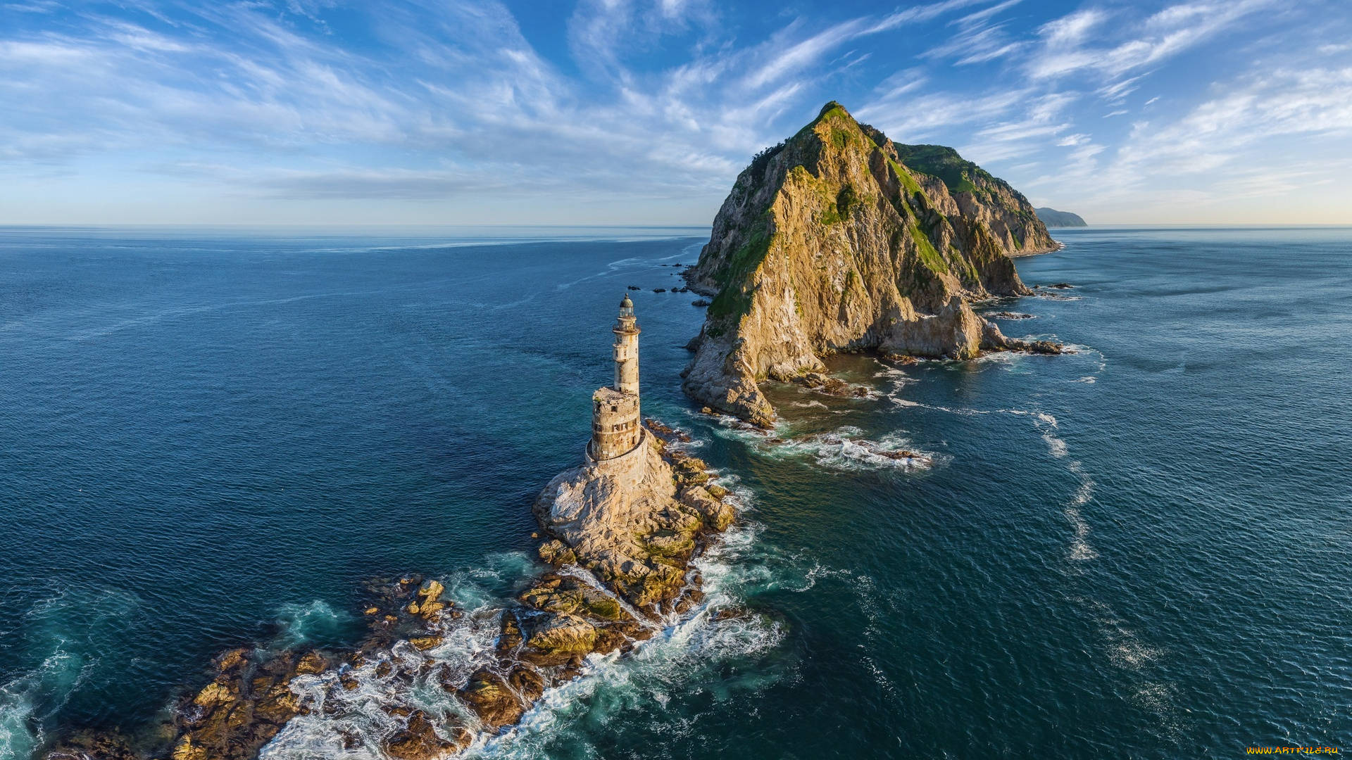 俄羅斯薩哈林島 壁紙 — 一座雄偉的石燈塔傲然矗立在崎嶇的海岸岩石上，海浪拍打其底部，廣闊的藍天中飄著縷縷白雲。