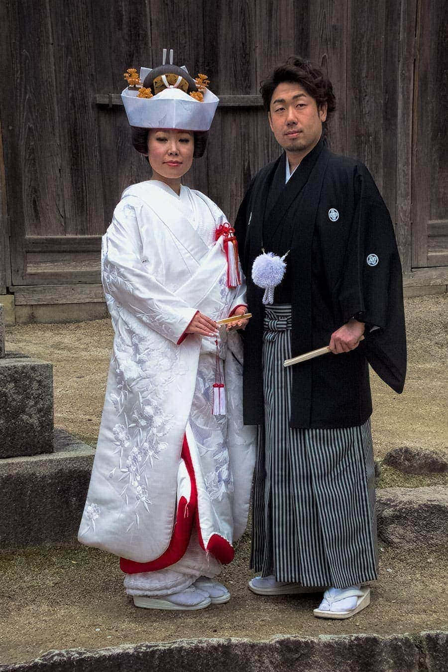 俏皮浪漫的花卉和服 壁紙 — 一對傳統日本新人穿著婚禮服飾，站在風化的木製神社門前。