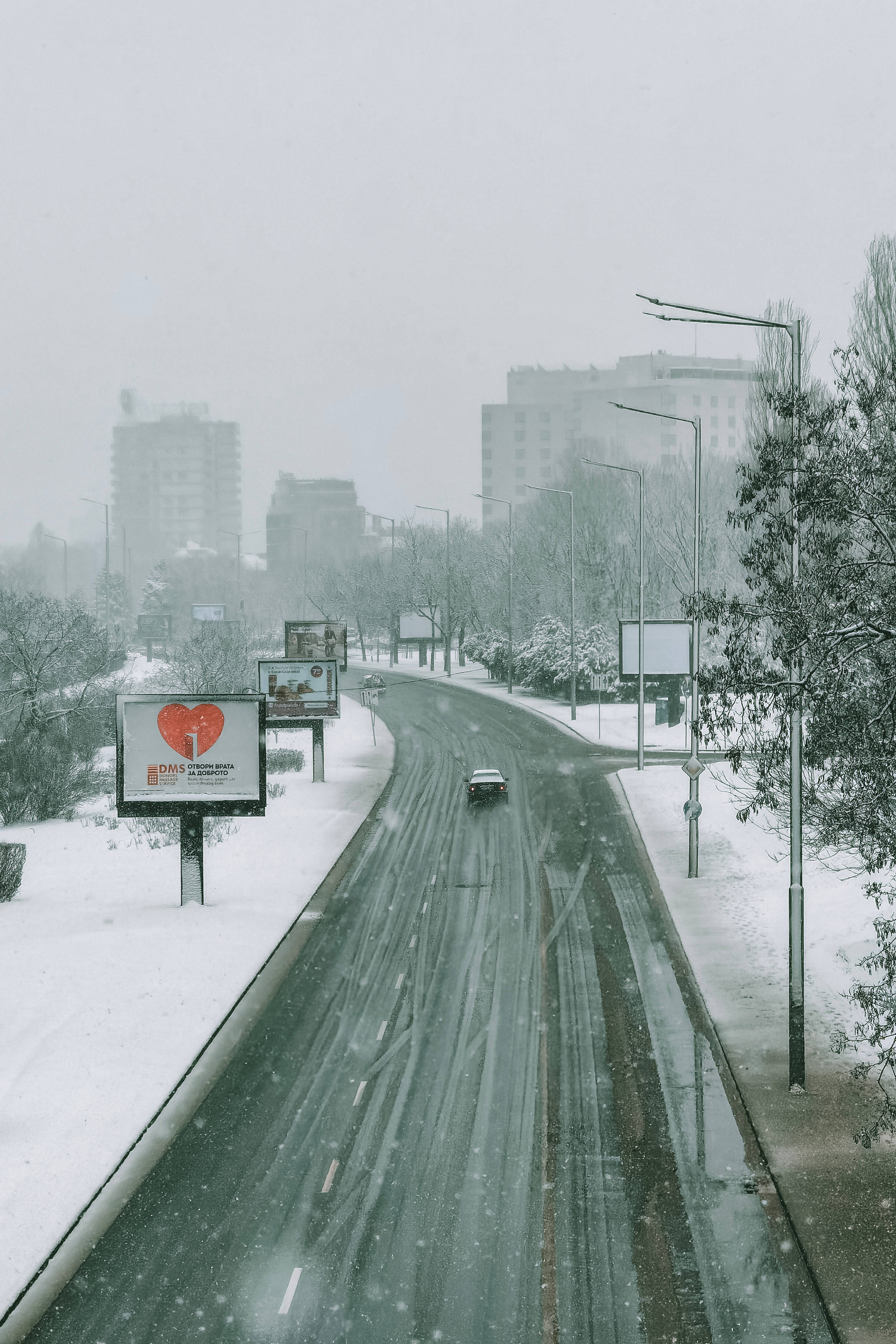 保加利亞冬季高速公路 壁紙 — 一輛孤獨的汽車在積雪的城市道路上行駛，大雪紛飛，建築物在灰色的冬日霧霾中逐漸消失。