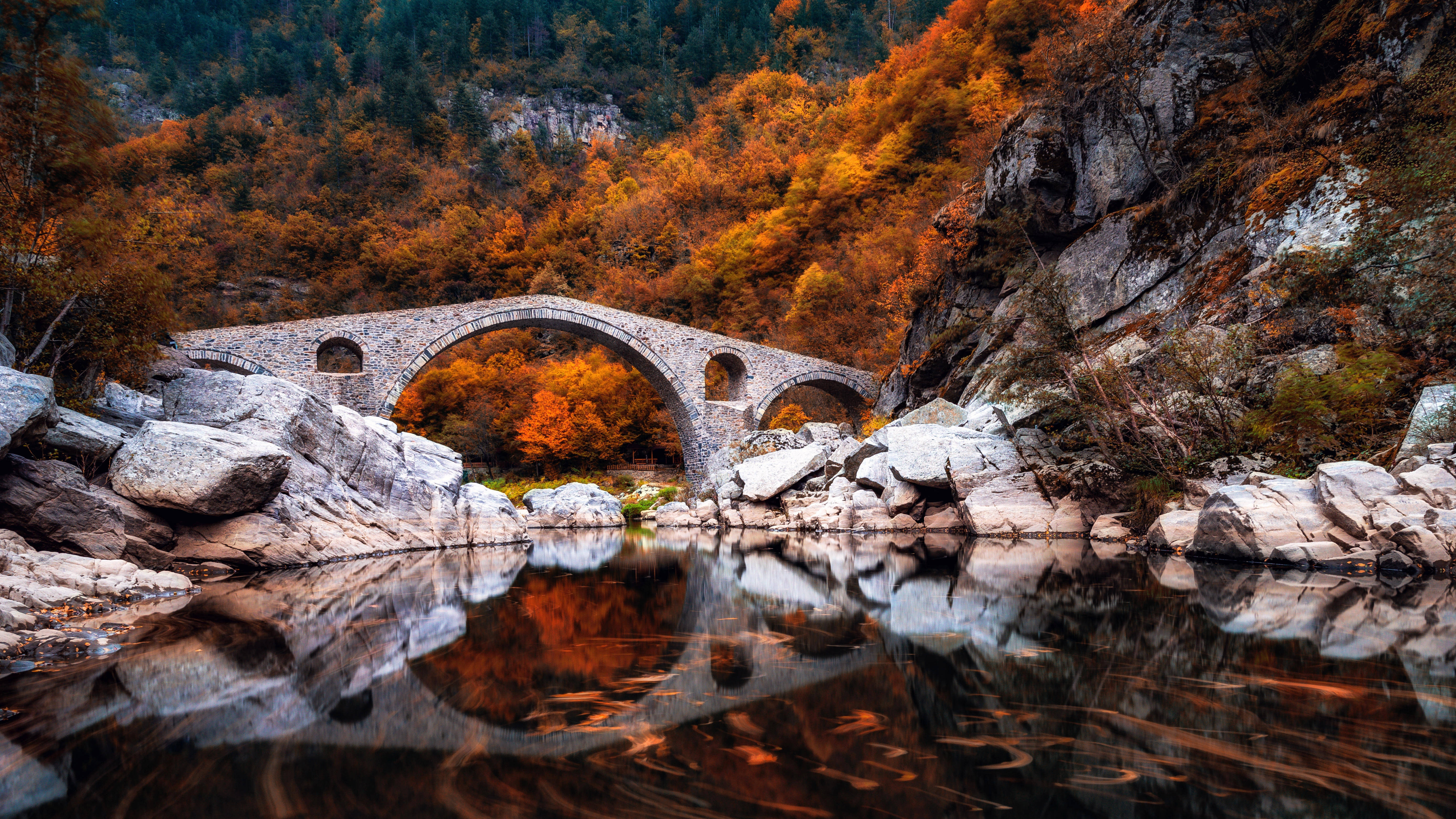 保加利亞魔鬼橋 壁紙 — 一座石拱橋橫跨平靜的河流，倒映著火紅的秋葉和崎嶇的懸崖，在柔和的光線下。