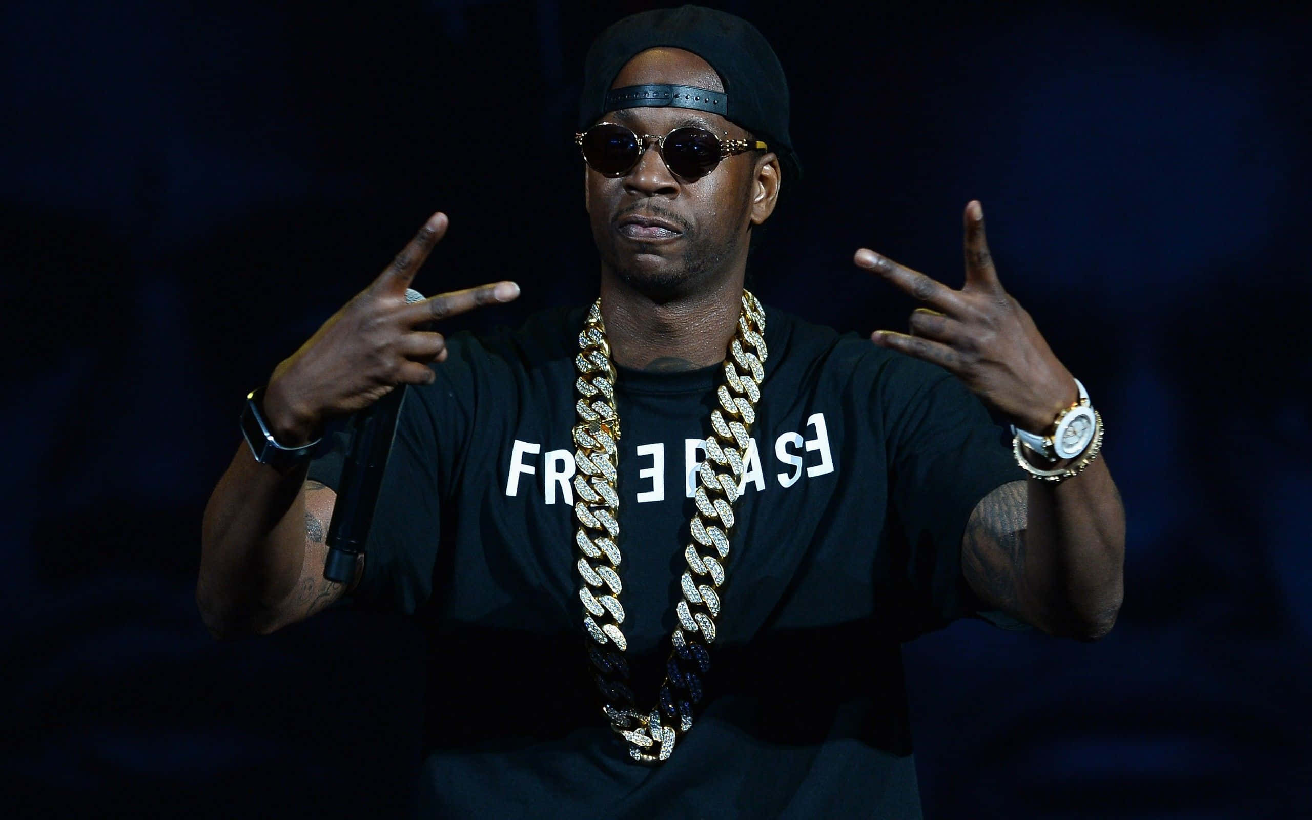 保持 2 Chainz 風格 壁紙 — 一位戴黑色帽和太陽眼鏡嘅饒舌歌手，喺舞台燈光下戴住金鏈同黑色T恤做手勢。