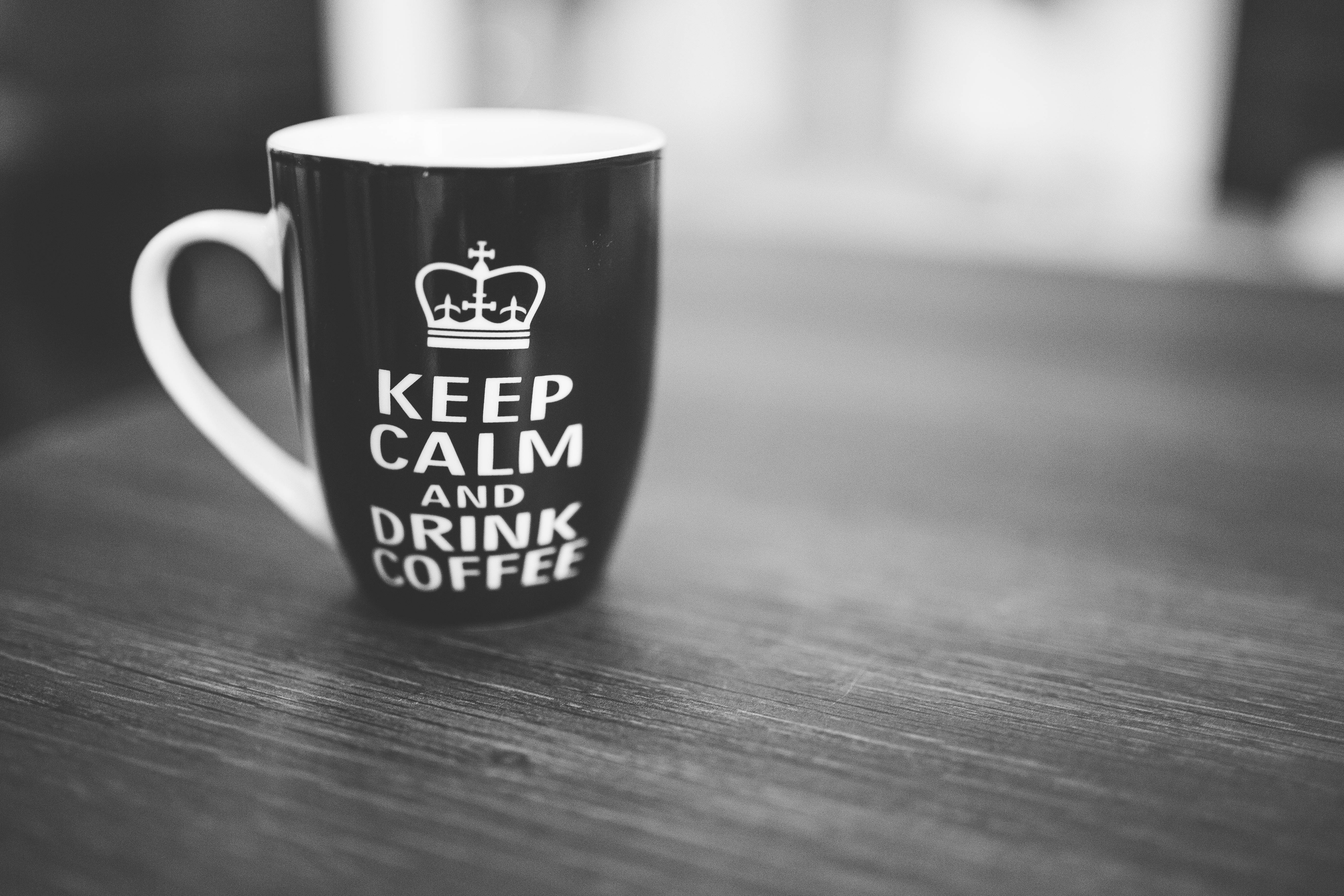 保持冷靜並喝咖啡 iPhone 壁紙 — 一個黑白馬克杯，上面有皇冠和「KEEP CALM AND DRINK COFFEE」字樣，放在木桌上。
