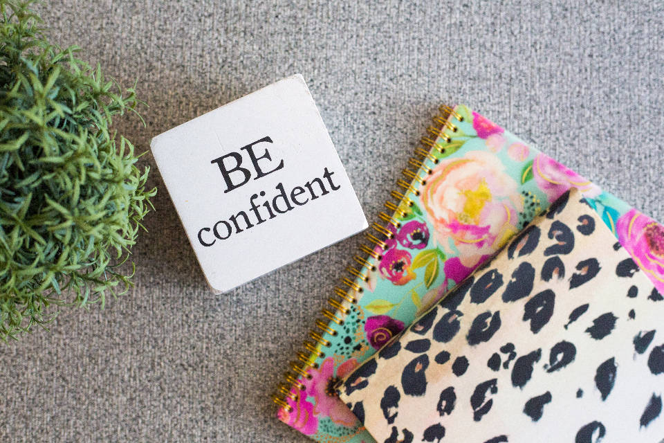 一個勵志方塊，上面寫著「BE confident」，旁邊有圖案筆記本和一棵綠色植物。