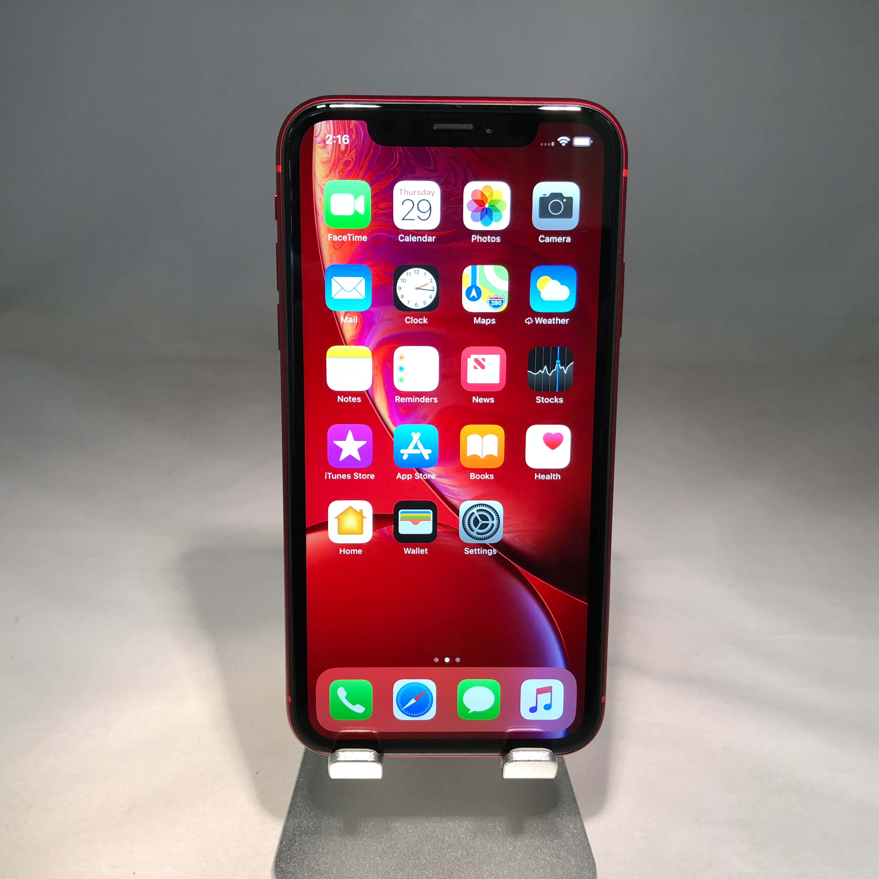 保持領先，掌握最新科技 壁紙 — 一部紅色iPhone XR直立於支架上，顯示主畫面，色彩繽紛的應用程式圖標襯托著旋轉的紅色牆紙。