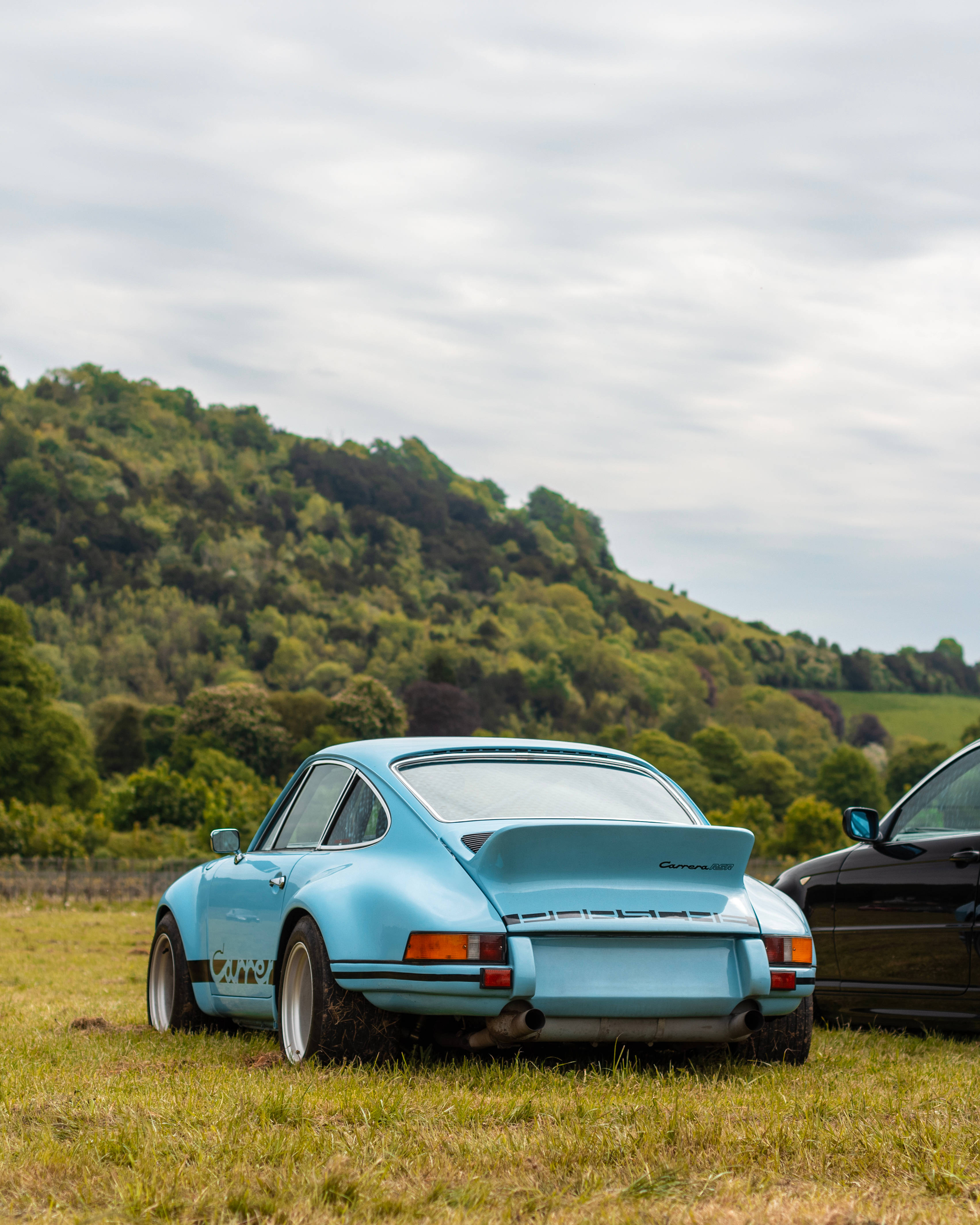 保時捷Carrera RSR豪華跑車 壁紙 — 一輛淺藍色經典保時捷911 Carrera RS停泊在草地上，背景是森林覆蓋的山丘，天空多雲。
