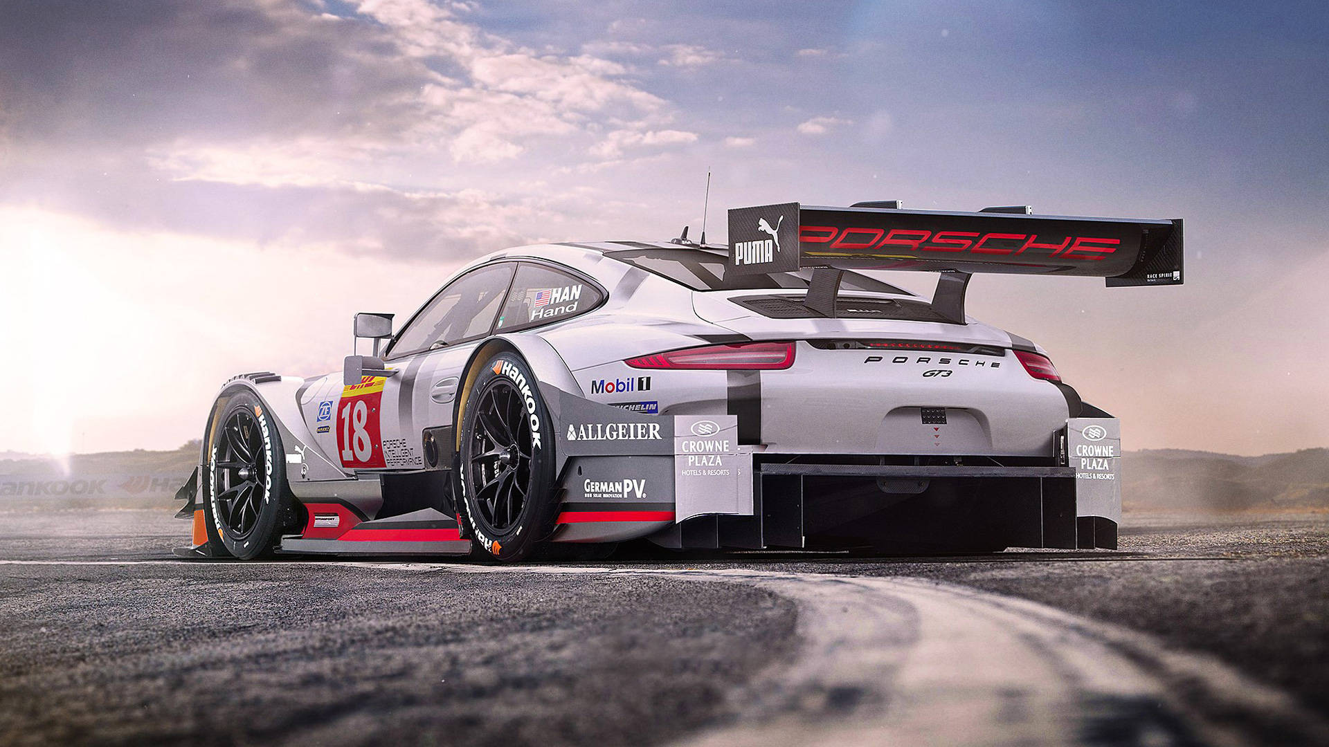 保時捷賽車競速 壁紙 — 一輛銀色保時捷911 GT3 R賽車，編號18，在戲劇性的日出天空下，沿著蜿蜒的瀝青賽道飛馳。