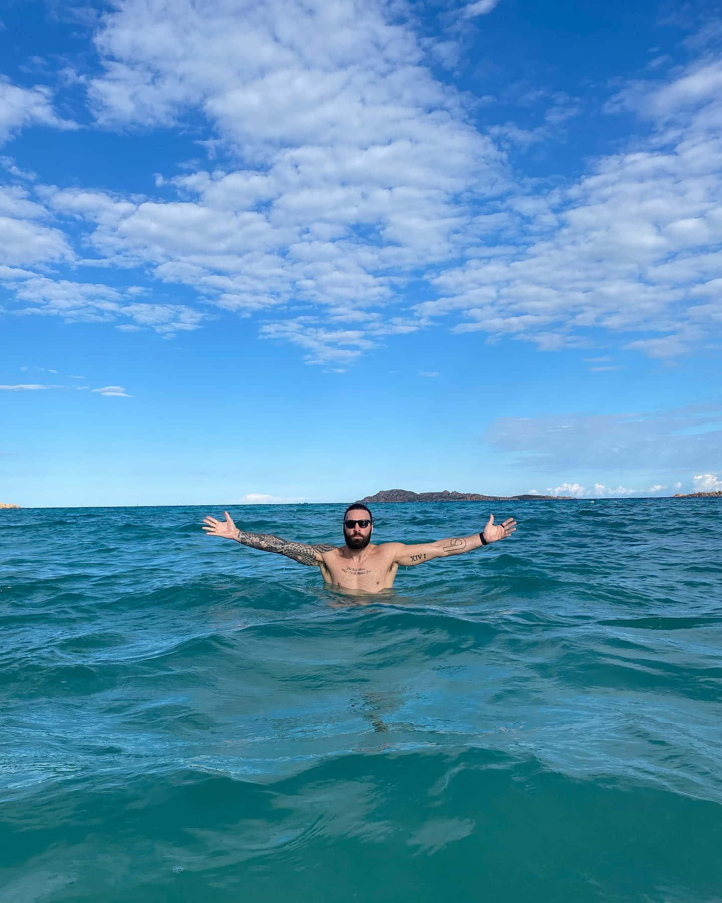 有紋身和太陽眼鏡的男人在藍天下於海水中張開雙臂