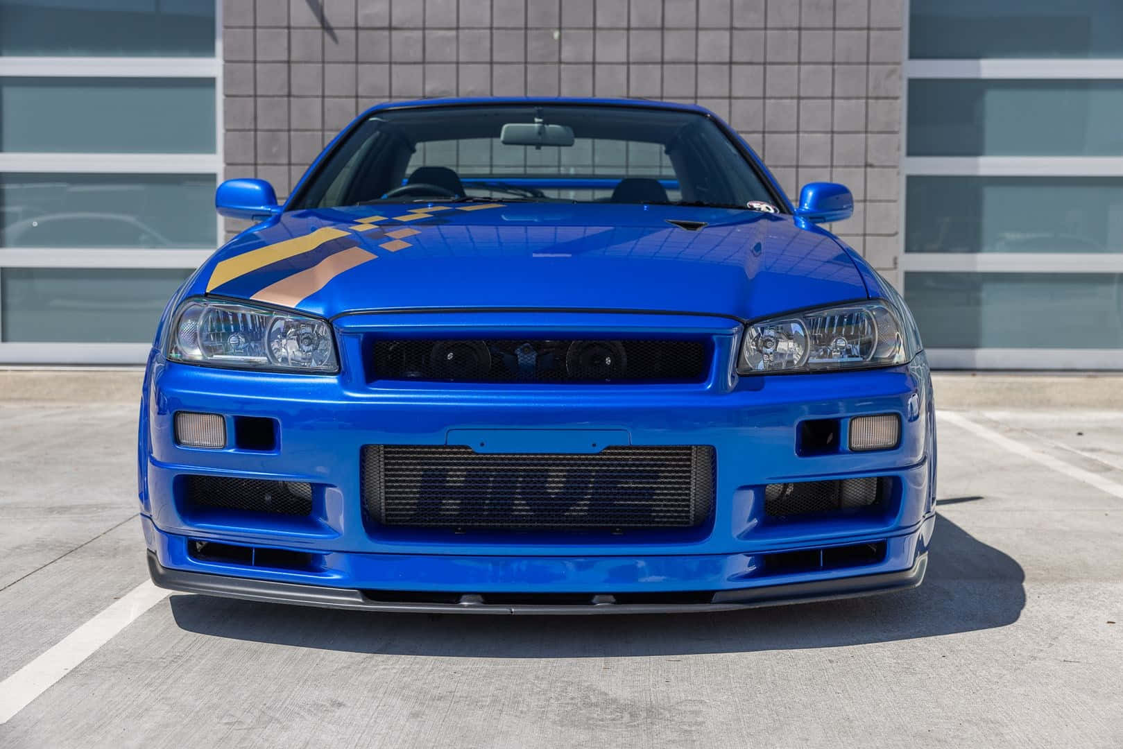 帶有賽車條紋的藍色日產Skyline R34停在一棟現代建築前。