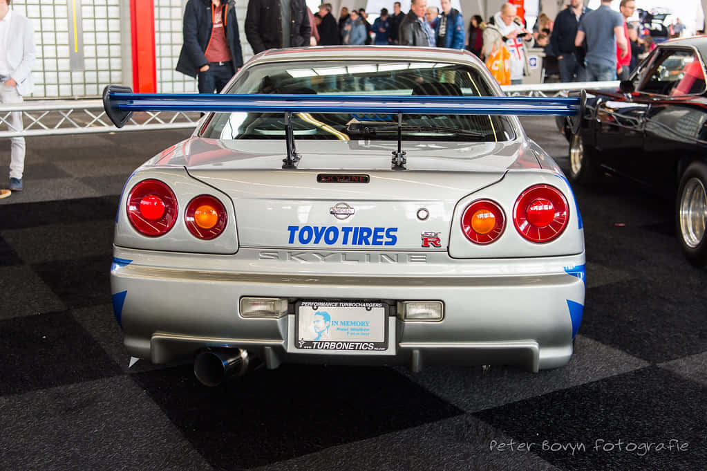 銀色日產Skyline R34，配備藍色尾翼，於室內車展中。