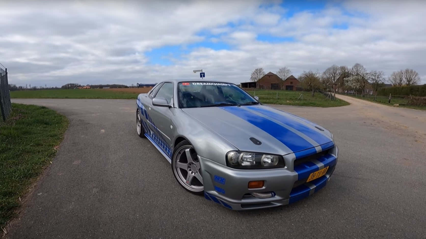 銀色日產Skyline R34，帶有藍色賽車條紋，停在鄉村道路上。