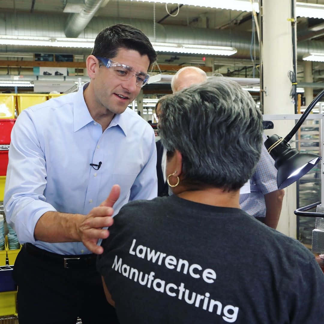 戴著安全眼鏡的政治人物與穿著 Lawrence Manufacturing 襯衫的工廠工人交談。