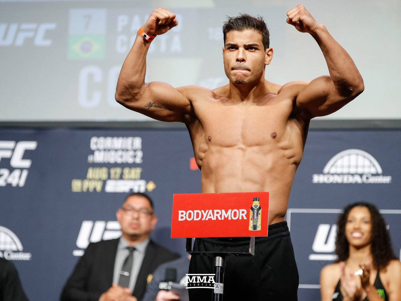 保羅·科斯塔，UFC中量級戰士，稱重時的英姿 壁紙 — 一位肌肉發達的UFC拳手在稱重活動中，於有品牌橫幅和官員的背景前，在磅秤上彎曲手臂。