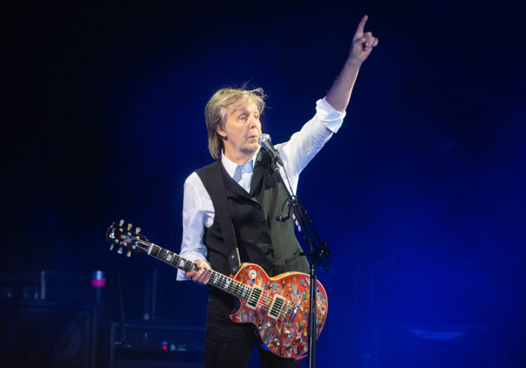 保羅·麥卡特尼演唱會指向上方 壁紙 — Paul McCartney 在舞台上表演，手持色彩繽紛的結他，在藍色舞台燈光下向上指。