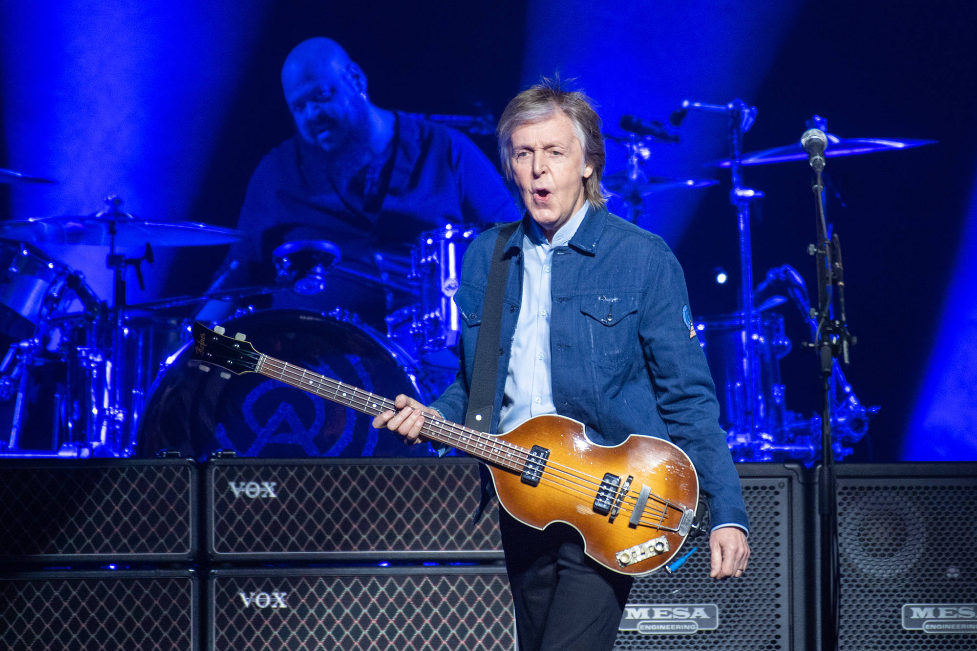 保羅·麥卡特尼演唱會搞笑嘴型 壁紙 — Paul McCartney 在充滿活力的藍色舞台燈光下熱情地彈奏一把漸變色貝斯結他，身後有一位鼓手。