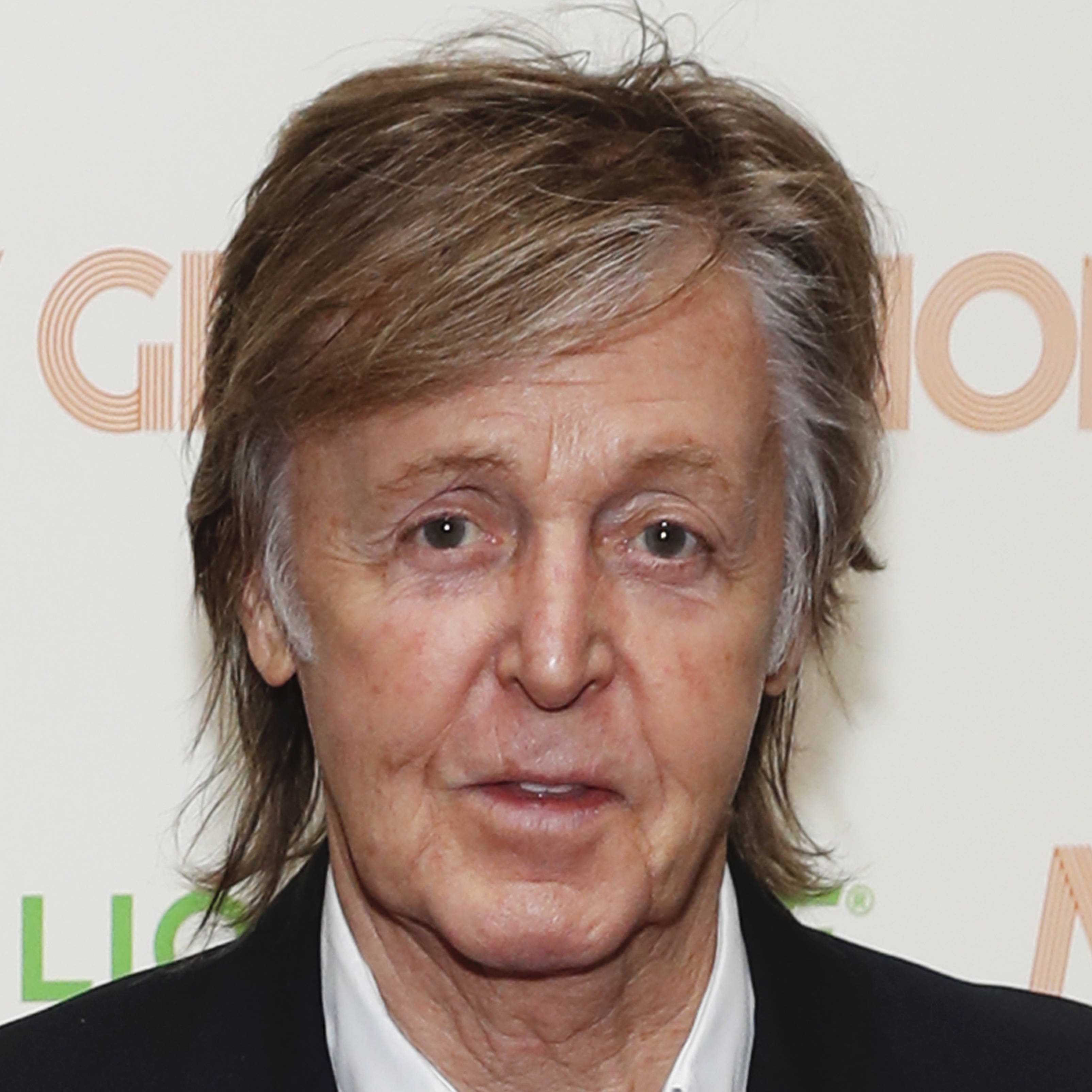 保羅·麥卡特尼顯老照片 壁紙 — Paul McCartney 嘅特寫肖像，佢有亂糟糟嘅棕色頭髮同灰色髮絲，著住黑色西裝同白色恤衫，背景係有品牌標誌嘅。