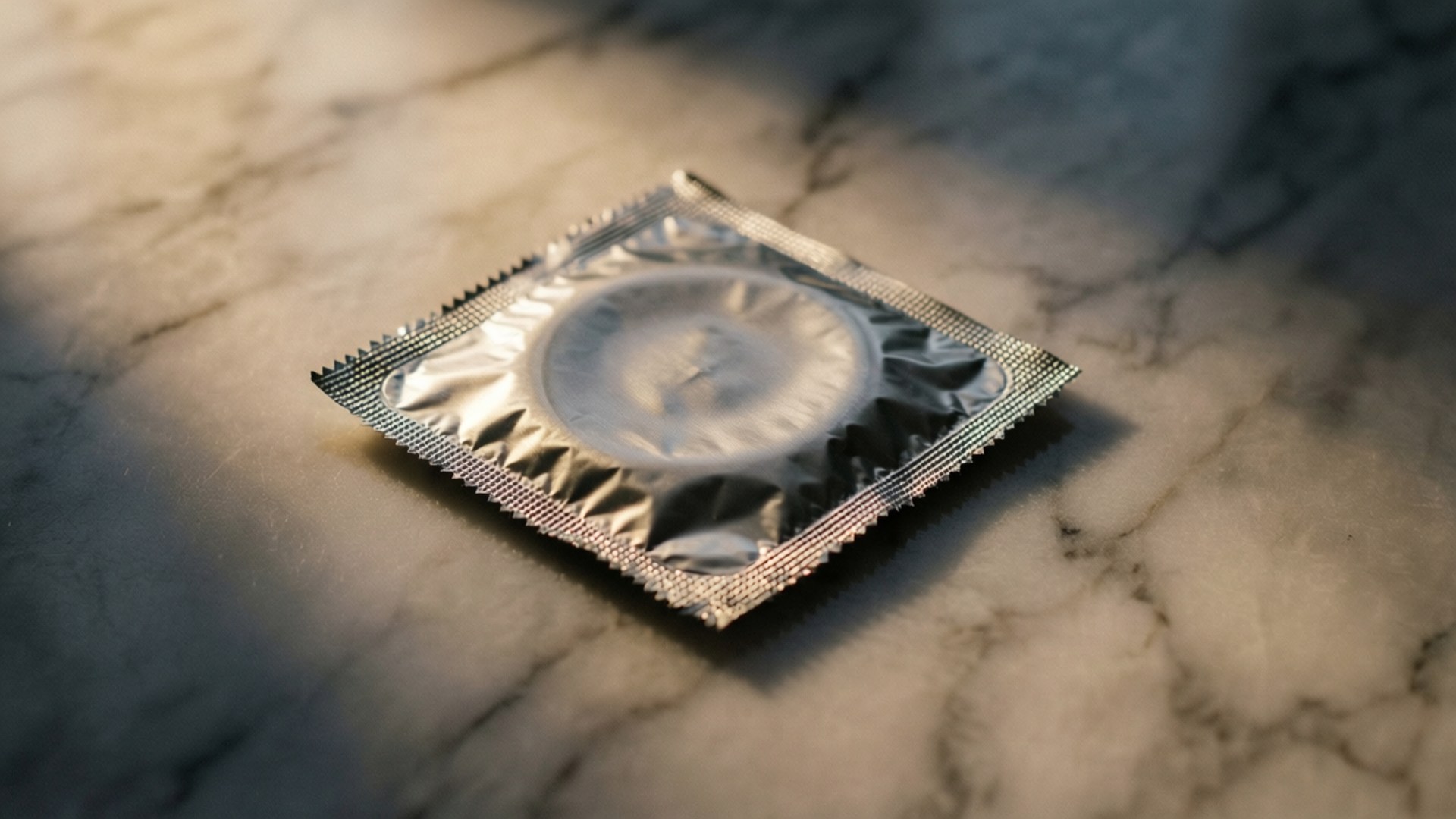 保險套優雅 壁紙 — 一個銀色錫紙安全套包裝放在大理石表面上，在柔和的方向性燈光下。