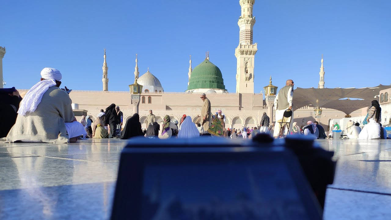 Madina Sharif 桌布 壁紙 — 信徒在陽光普照的大理石庭院中，在藍天白雲下，於一座擁有綠色圓頂和宣禮塔的宏偉清真寺前祈禱。
