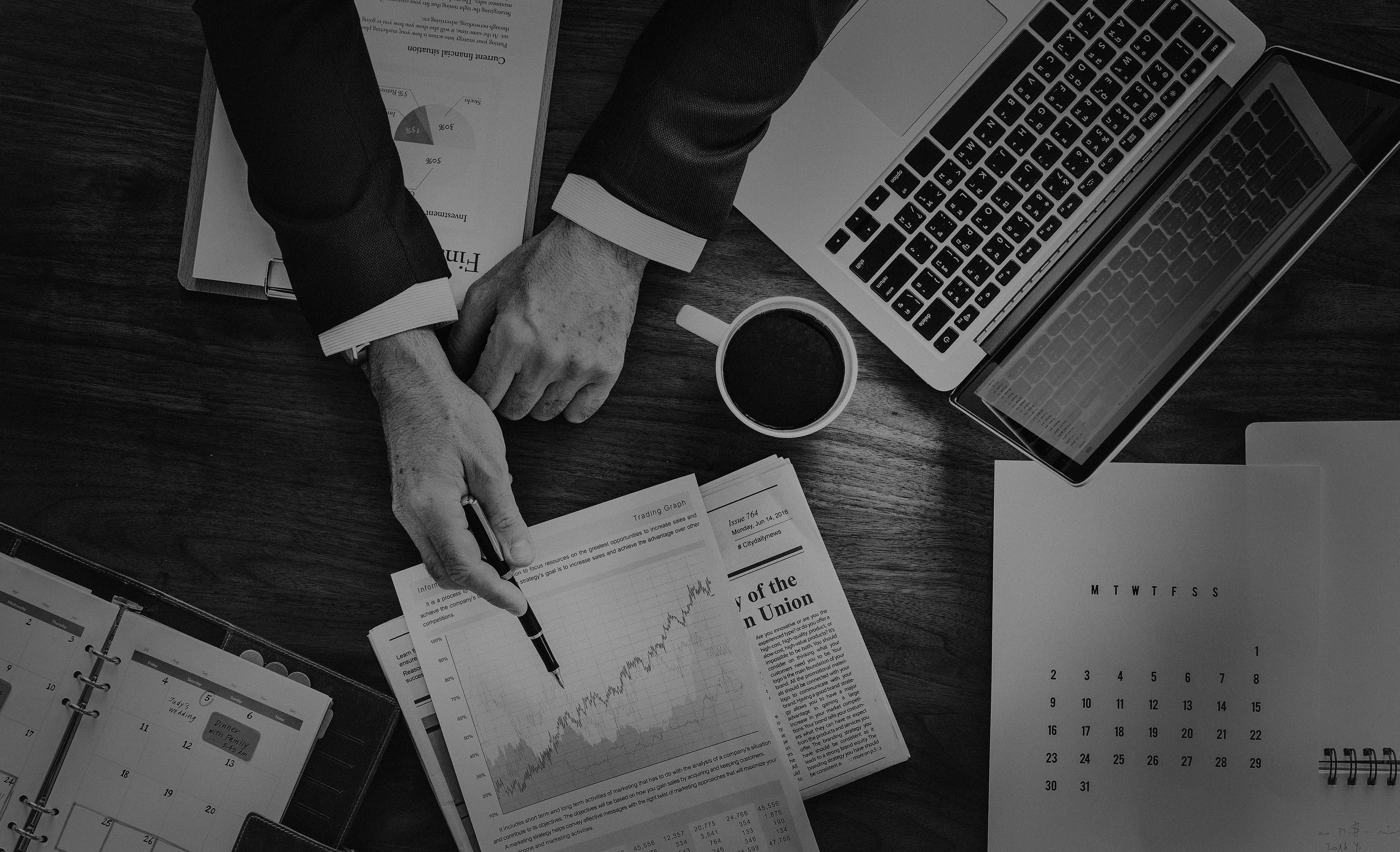 Business 桌布 壁紙 — 俯拍黑白照片，一名商人的雙手在木桌上分析金融圖表和圖形，桌上放有手提電腦和咖啡杯。