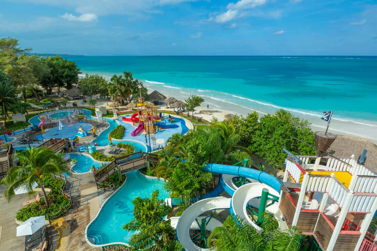 Jamaican Beach 桌布 壁紙 — 俯瞰一個充滿活力的熱帶度假村，擁有碧綠的泳池、水上滑梯和棕櫚樹，可欣賞純淨的海灘和海洋。