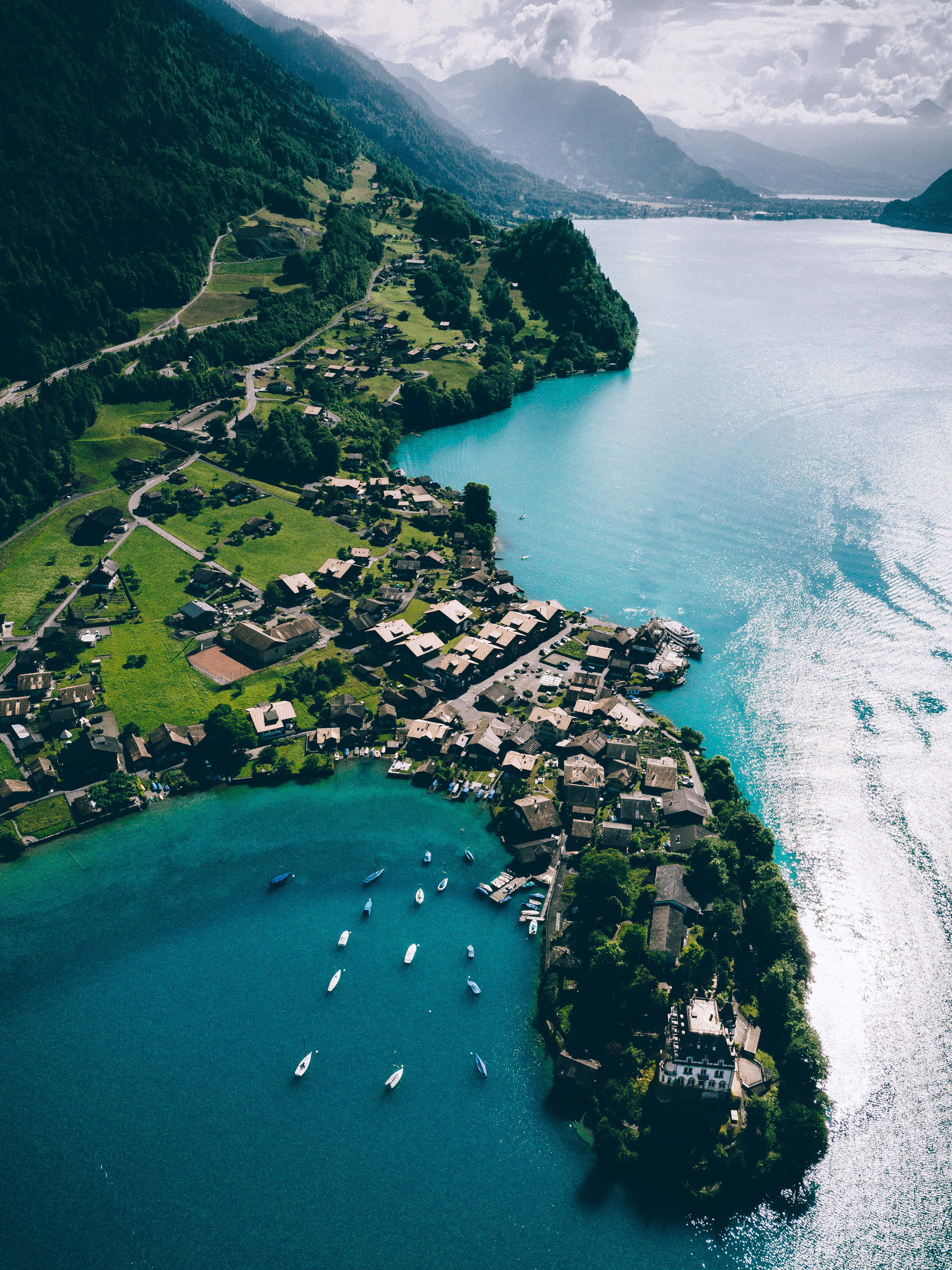 Switzerland 桌布 壁紙 — 俯瞰一個古色古香的湖邊村莊，坐落在鬱鬱蔥蔥的綠色山丘中，旁邊是一個充滿活力的碧綠湖泊，點綴著船隻。