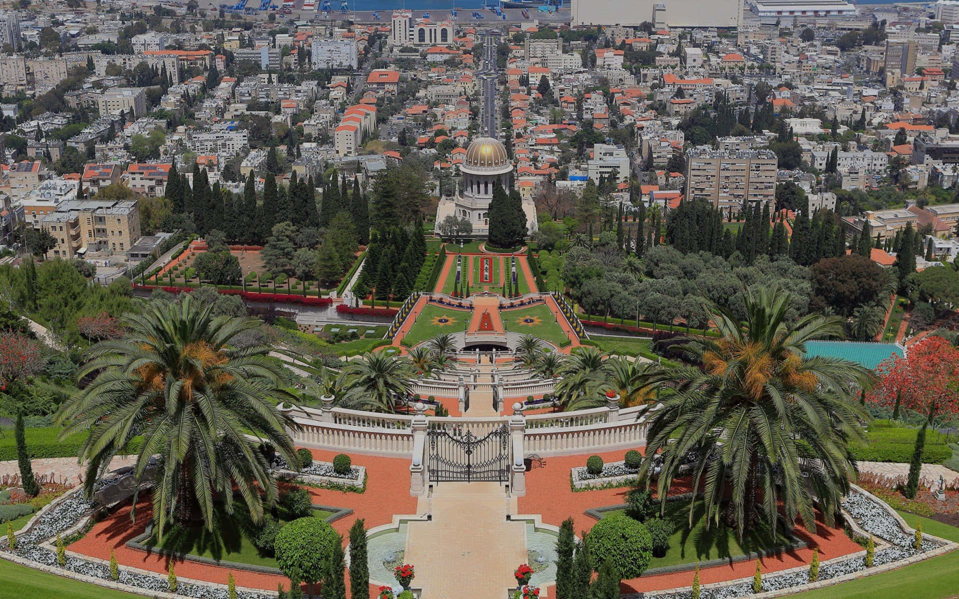 Israel 桌布 壁紙 — 俯瞰一個宏偉花園的空中景觀，花園內有棕櫚樹和一座金色圓頂建築，俯瞰著密集的城市景觀。