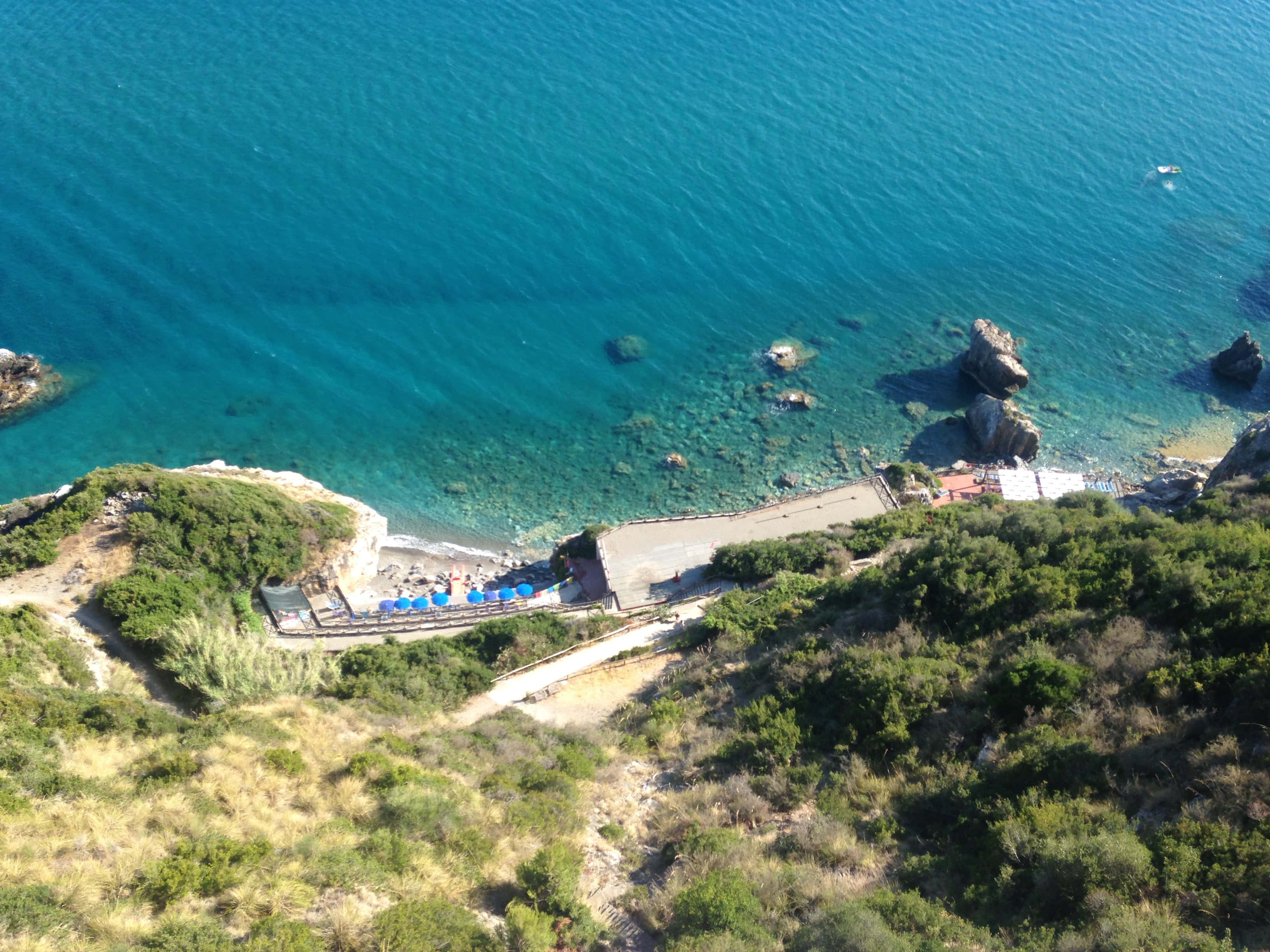 Italian Beach 桌布 壁紙 — 俯瞰一個隱蔽的藍綠色海灣，擁有岩石海岸和藍色遮陽傘，排列在一個被茂密綠色山丘環抱的海灘上。