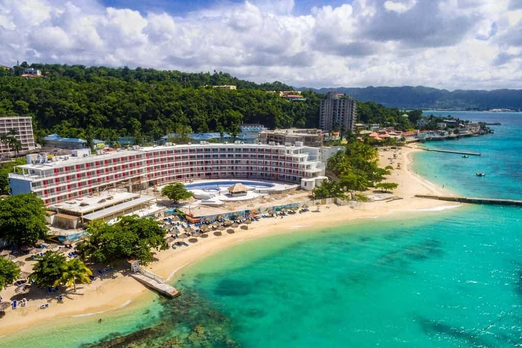 Jamaican Beach 桌布 壁紙 — 俯瞰一座弧形度假酒店，坐落在沙灘上，周圍有碧綠的海水和茂密的綠色山丘，天空部分多雲。