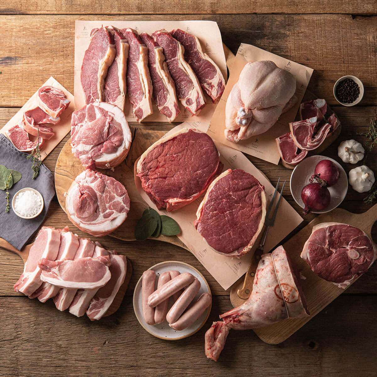 Red Meat 桌布 壁紙 — 俯瞰各類生肉，包括牛扒、豬扒、香腸及整塊切肉，排列在木板上，配以大蒜、洋蔥及香草，置於鄉村風木桌上。