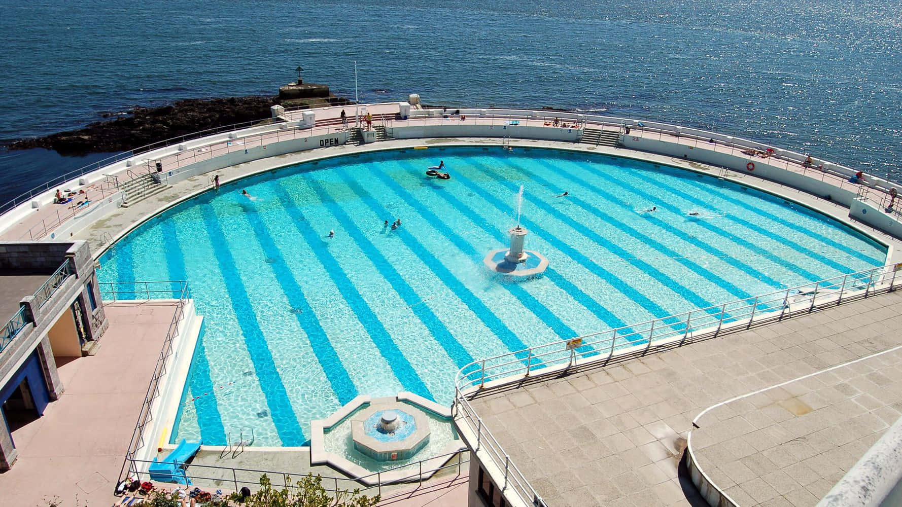 Beach Swimming Pool 桌布 壁紙 — 俯瞰彎曲的海濱泳池，泳客在條紋藍色海水中暢泳，旁邊是岩石海岸線。