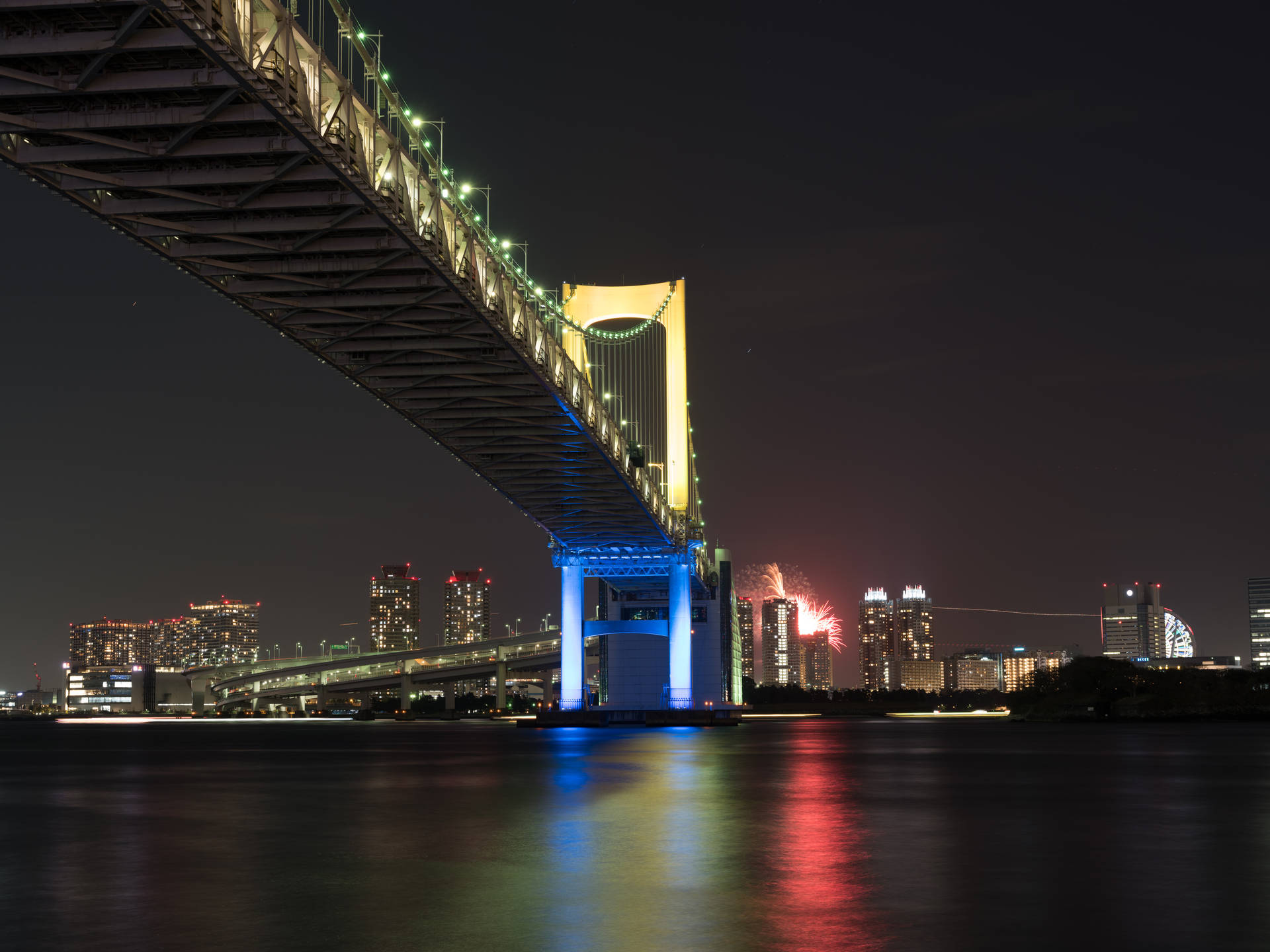 彩虹橋在夜間亮起，煙火在東京天際線上綻放，倒映在水面上。