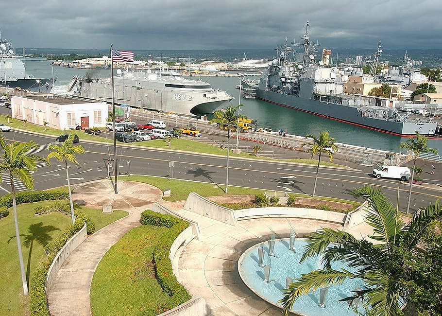 俯瞰珍珠港港口 壁紙 — 鳥瞰一個海軍基地，大型灰色軍艦停泊在港灣，周圍有棕櫚樹和城市基礎設施，天空多雲。
