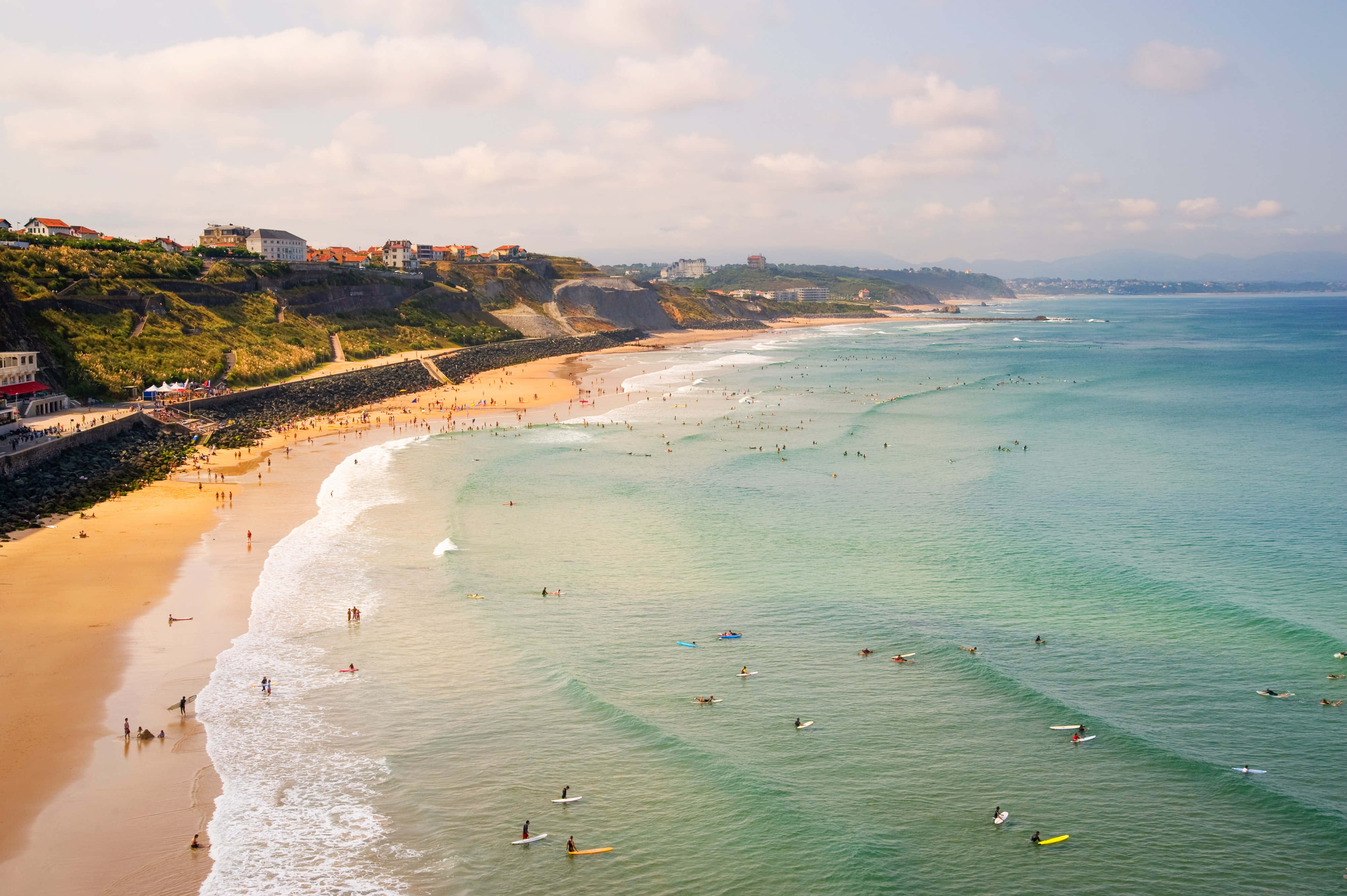 French Beach 桌布 壁紙 — 俯瞰繁忙海灘，在部分多雲的天空下，碧綠海水和金色沙灘上有衝浪者。