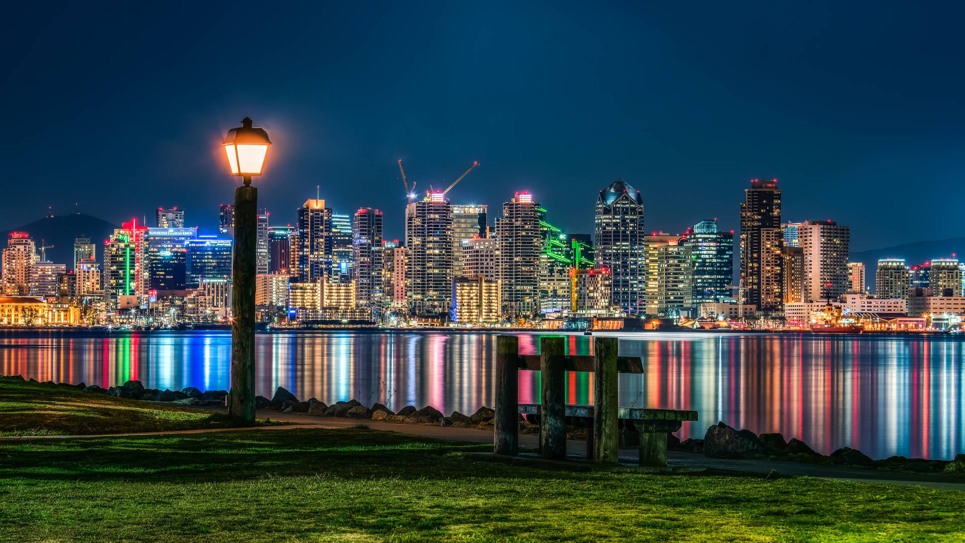城市天際線在水中的倒影夜景，前景有一盞亮著的街燈和長椅。