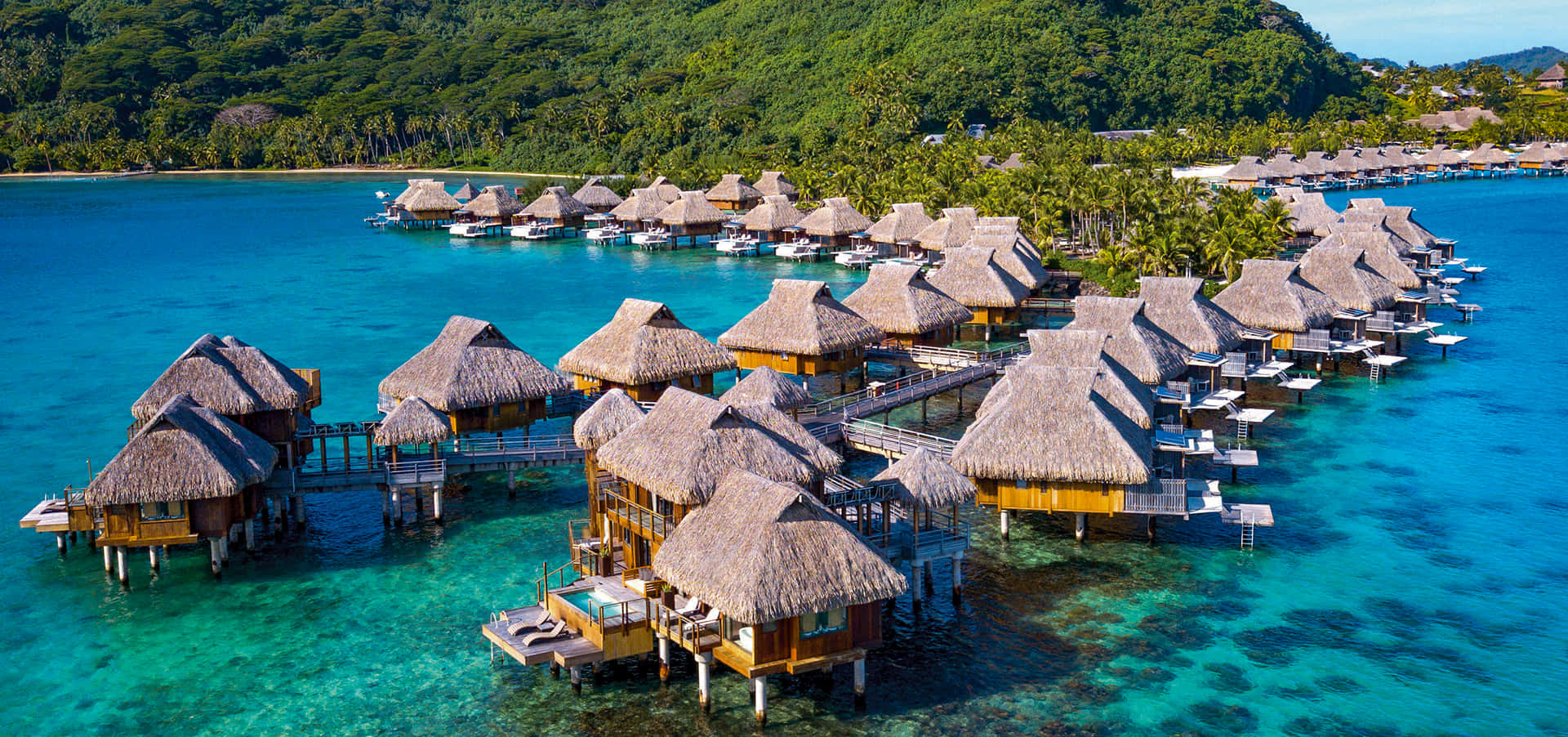 Bora Bora Beach 桌布 壁紙 — 俯瞰茅草屋頂水上平房，坐落在晶瑩剔透的碧綠海水旁，毗鄰鬱鬱蔥蔥的熱帶島嶼。