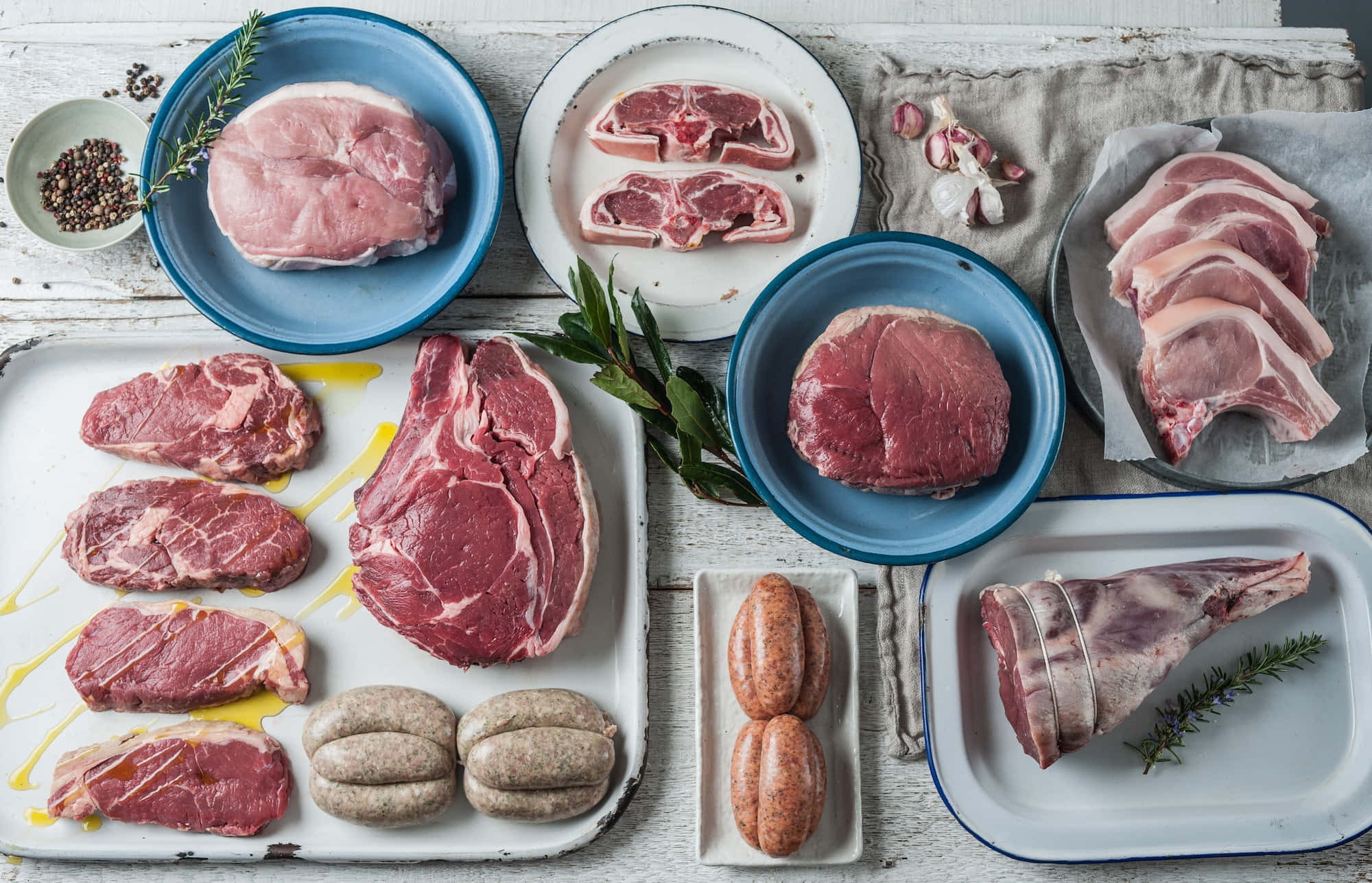 Red Meat 桌布 壁紙 — 俯瞰鏡頭下，各種生肉包括豬扒、牛扒、香腸和羊腿，放在盤子上，配上香草和油，置於鄉村風格的木質表面上。