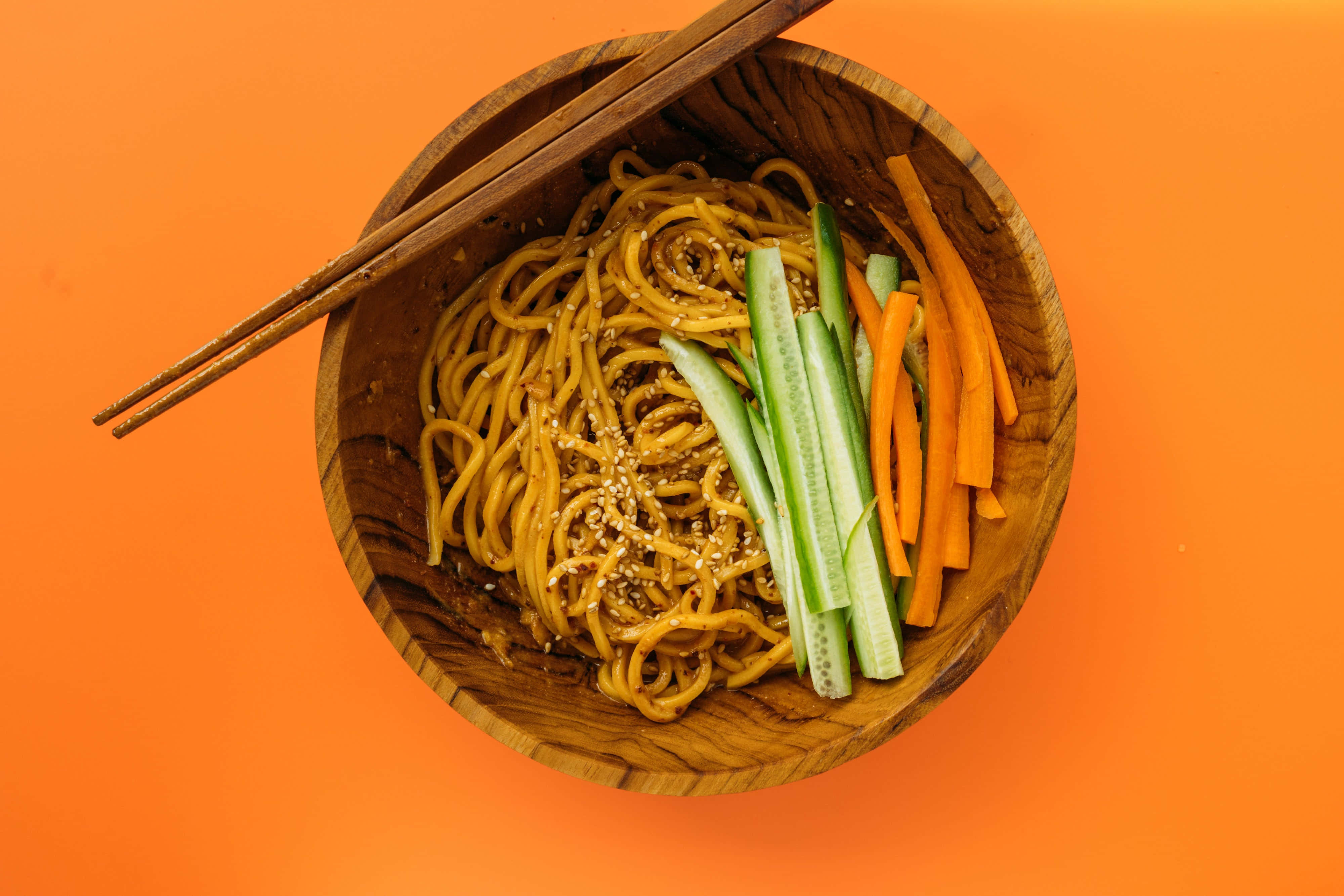 Noodles 桌布 壁紙 — 俯視一個木碗，裡面裝滿了芝麻麵條和切片青瓜及紅蘿蔔，背景是鮮豔的橙色。